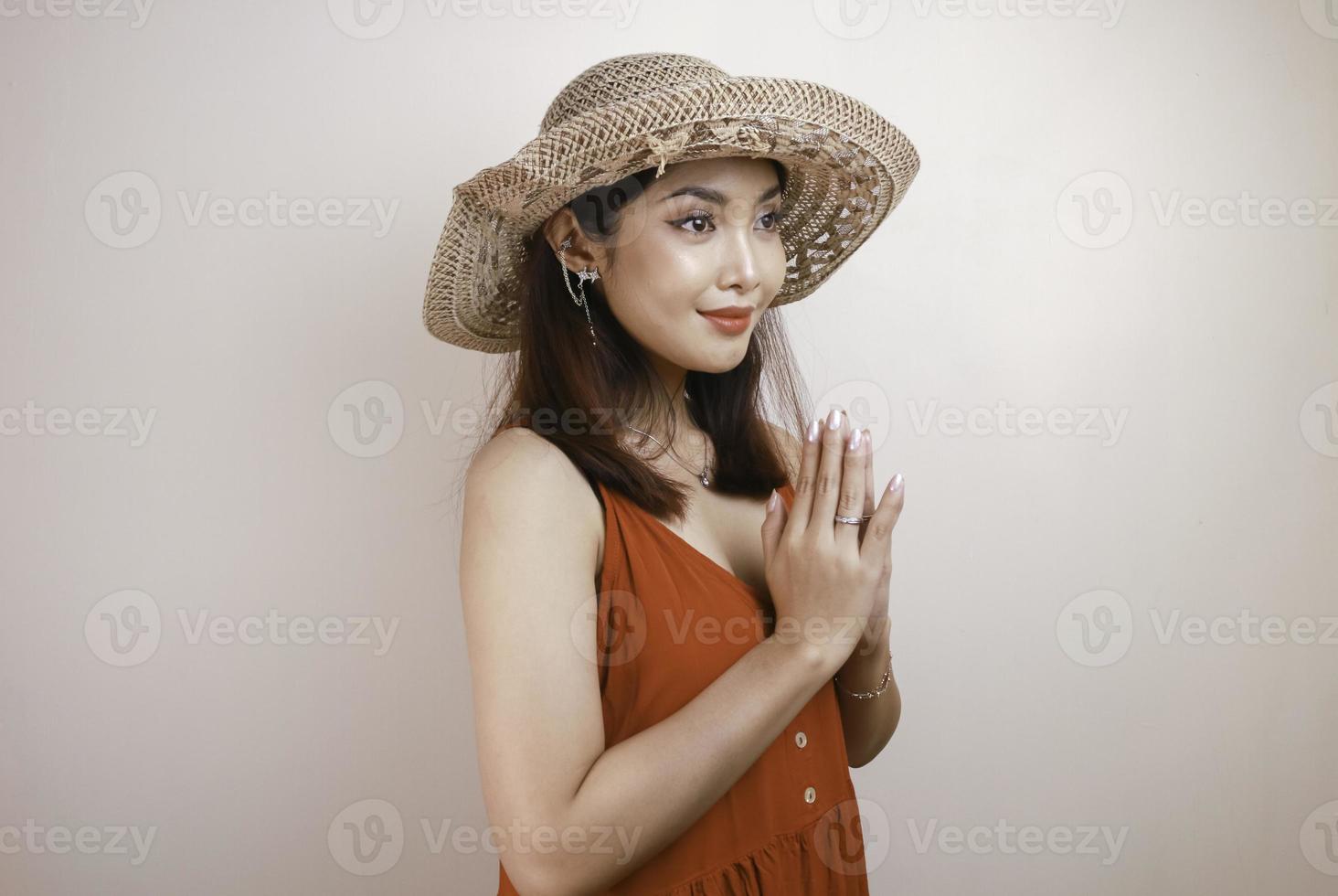 eine junge schöne asiatische frau, die einen strohhut trägt, gibt grüßende hände mit einem breiten lächeln auf ihrem gesicht. indonesische Frau auf weißem Hintergrund. foto