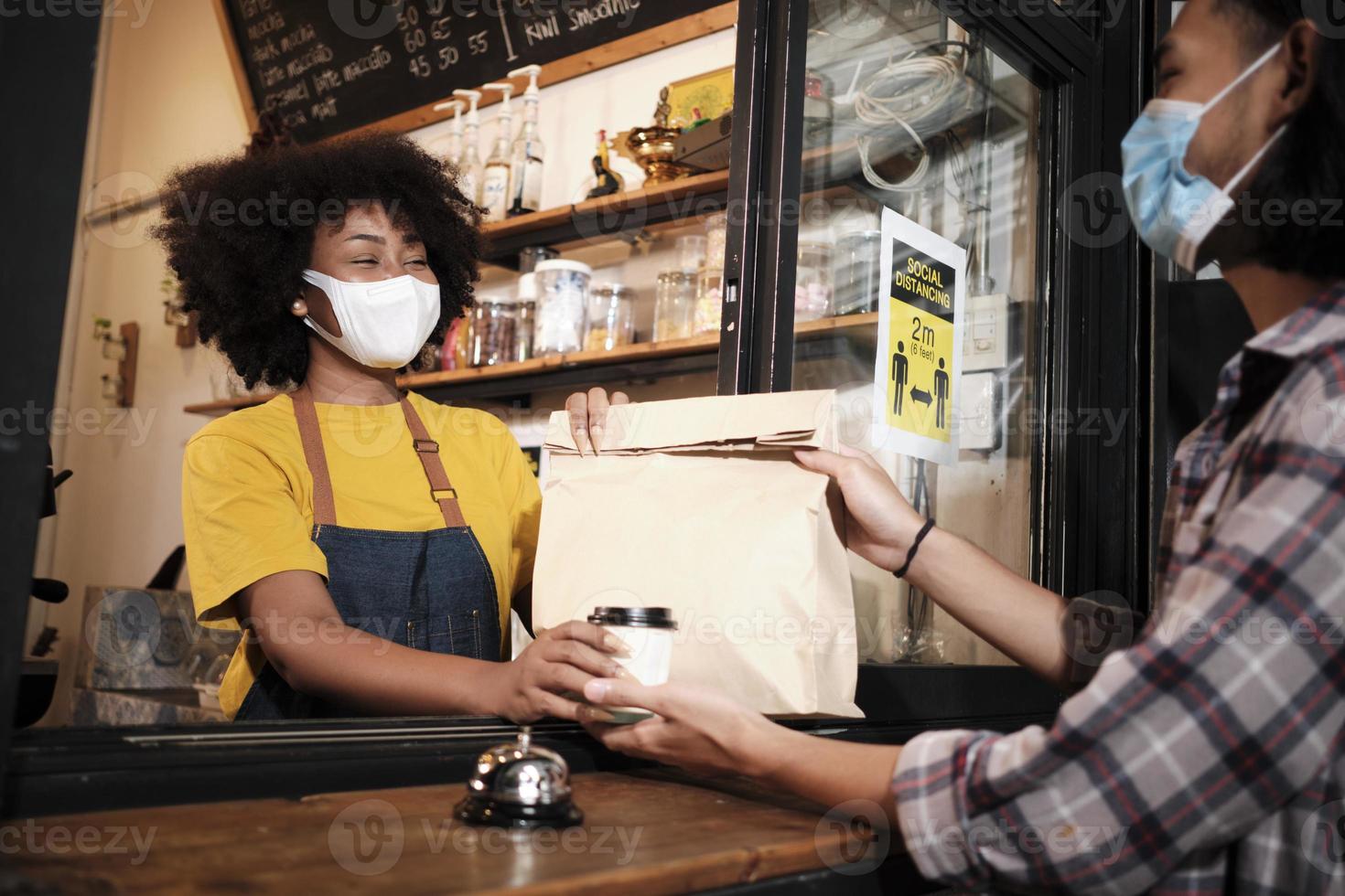 afroamerikanische barista mit gesichtsmaske arbeitet im café mit sozialer distanz, kaffee zum mitnehmen für einen kunden, neuer normaler service des cafés für kleine unternehmen im covid19-quarantänelebensstil. foto