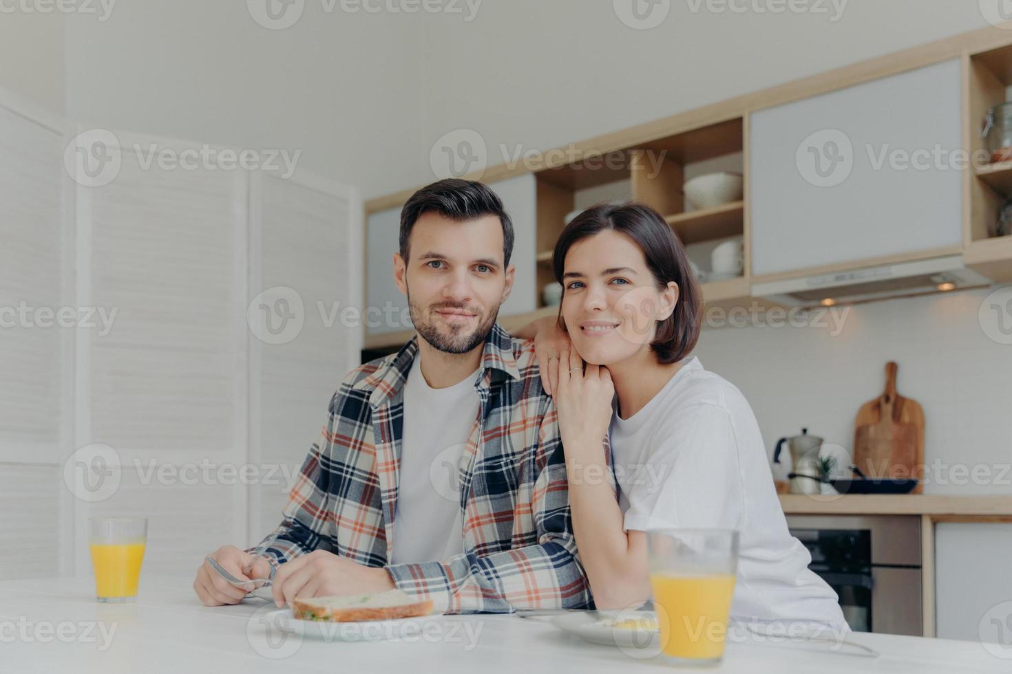 positive brünette frau lehnt sich an die schulter ihres mannes, posiert gemeinsam in der küche, genießt leckeres frühstück, hat fröhliche stimmung, schaut direkt in die kamera, hat gute beziehungen. Familienkonzept foto