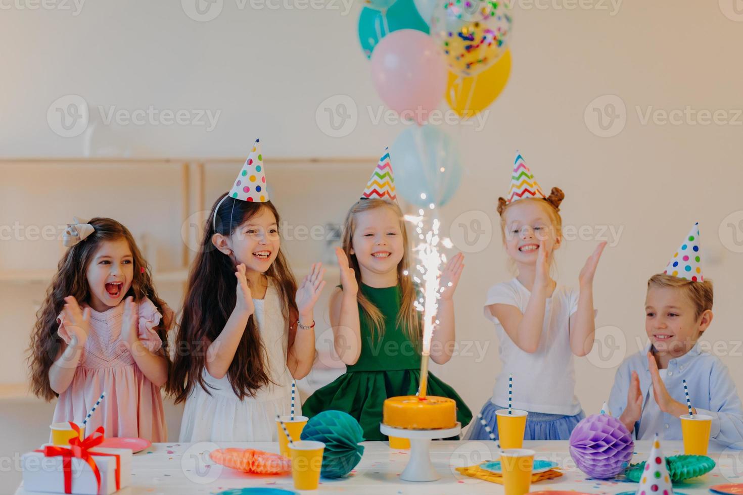 überglückliche kinderfreunde klatschen in die hände und schauen auf den kuchen, stehen in der nähe eines festlichen tisches mit geschenken, tassen und feiertagsattributen, haben gemeinsam spaß, feiern geburtstag foto