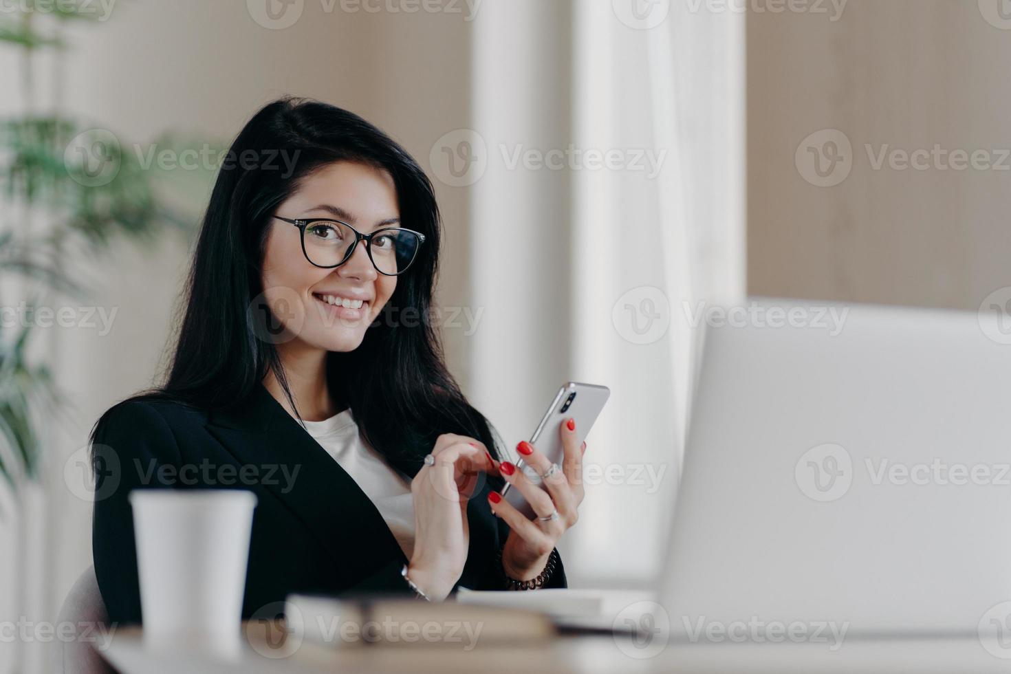 horizontale aufnahme von glücklichen brünetten europäischen frauennachrichten auf modernem smartphone, gibt online-beratung, lächelt positiv, trägt brille und formellen anzug, sitzt vor geöffnetem laptop. foto