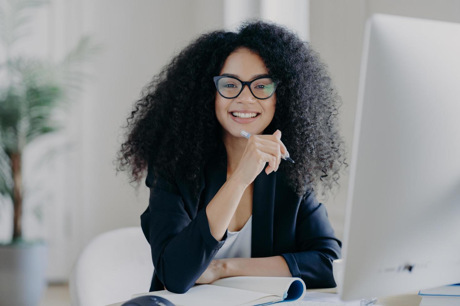 positive internationale studentin mit krausem haar, trägt durchsichtige brille, hält stift in der hand, macht buchhaltung, sitzt vor großem computerbildschirm, gekleidet in schwarzes elegantes outfit. foto