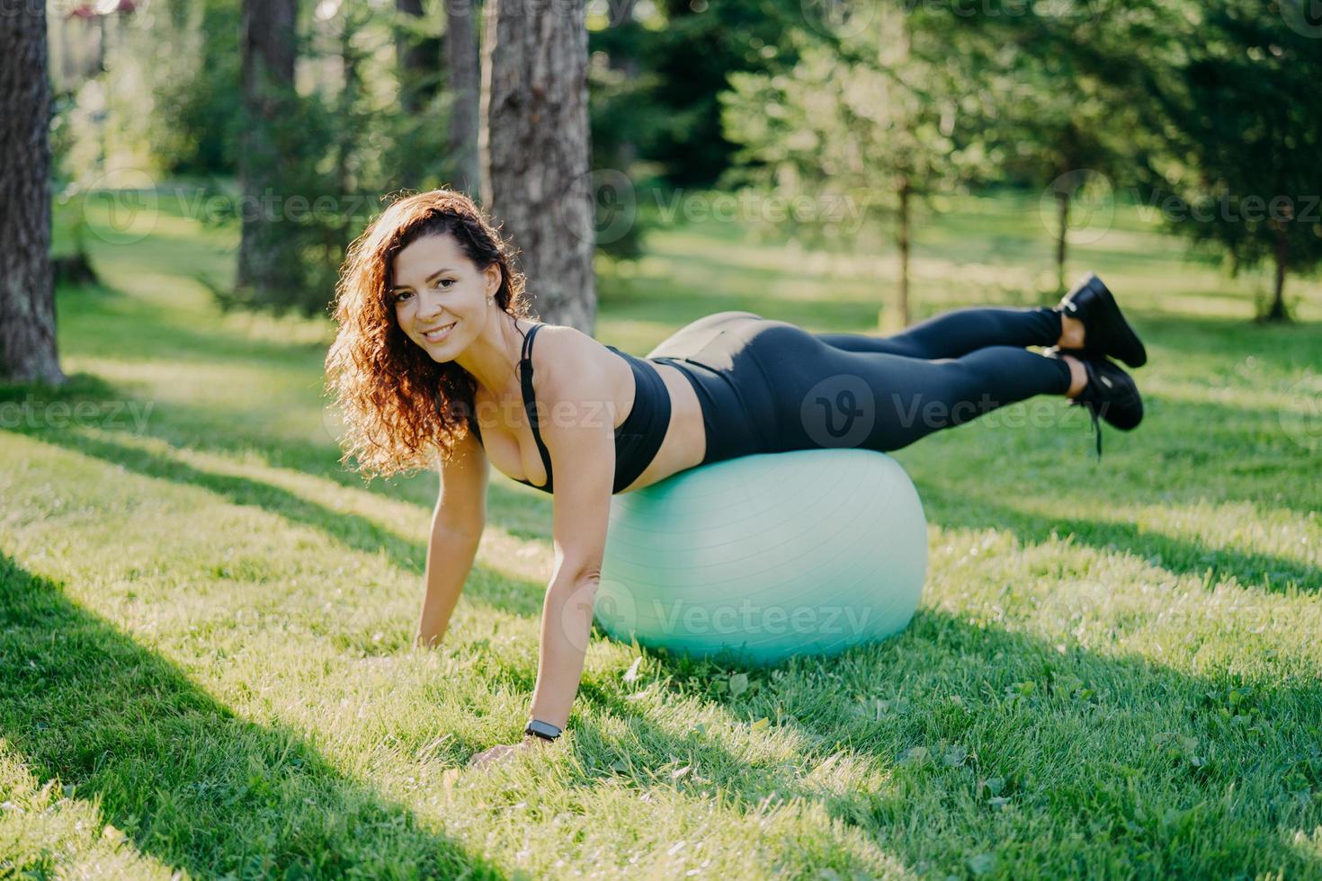 sport, erholung, fitness und gesundes lebensstilkonzept. brünette sportliche junge europäische frau in aktiver kleidung lehnt sich an fitnessball, macht körperliche übungen im freien, genießt lieblingsbeschäftigung foto