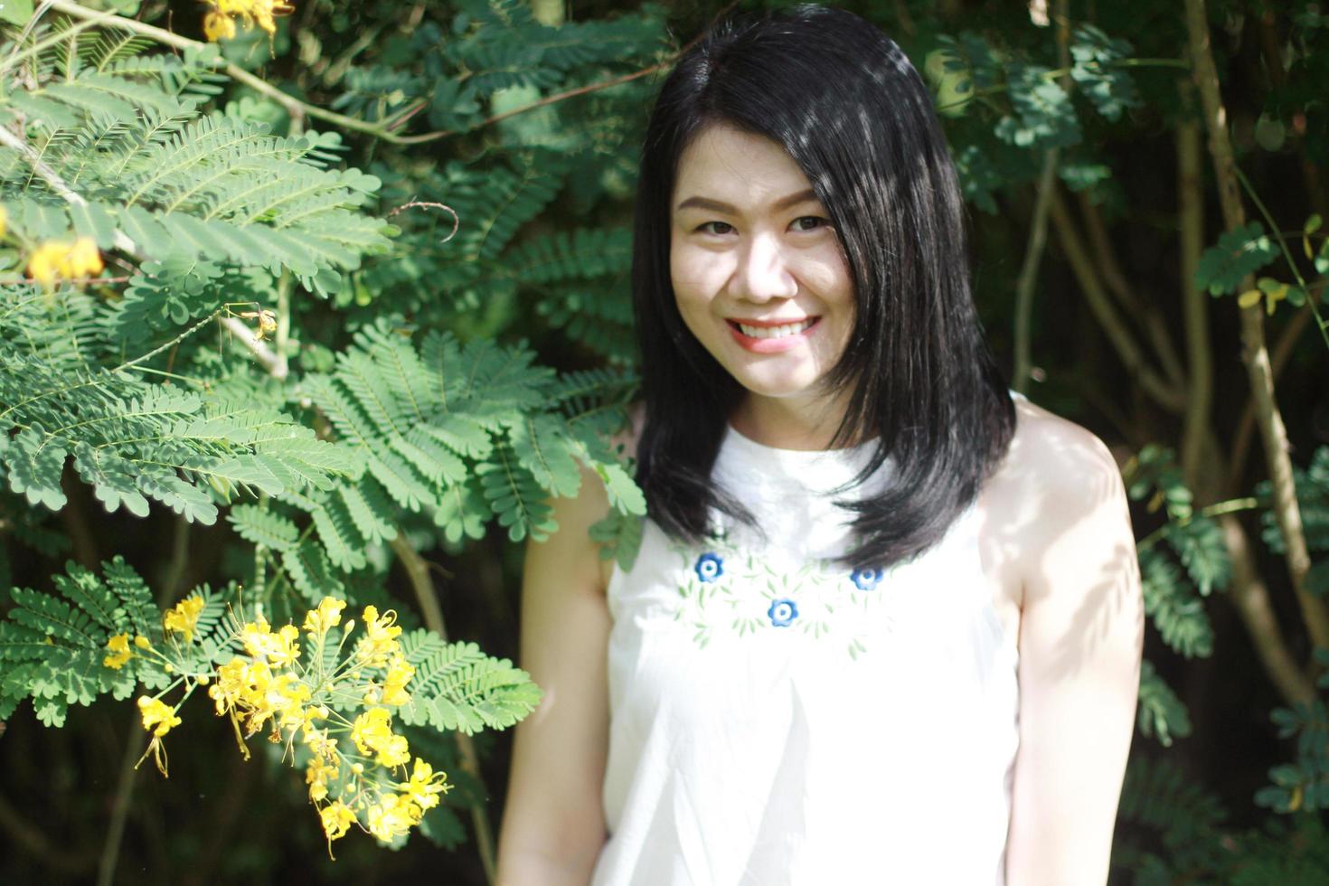 schöne asiatische frau im weißen kleid entspannen und lächeln im naturpark. junges thailändisches mädchen genießen im urlaub mit sonnenlicht im gelben blumengarten foto