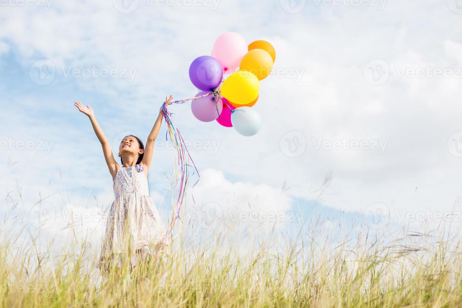 süßes kleines mädchen, das bunte luftballons auf der wiese gegen blauen himmel und wolken hält und die hände ausbreitet. foto