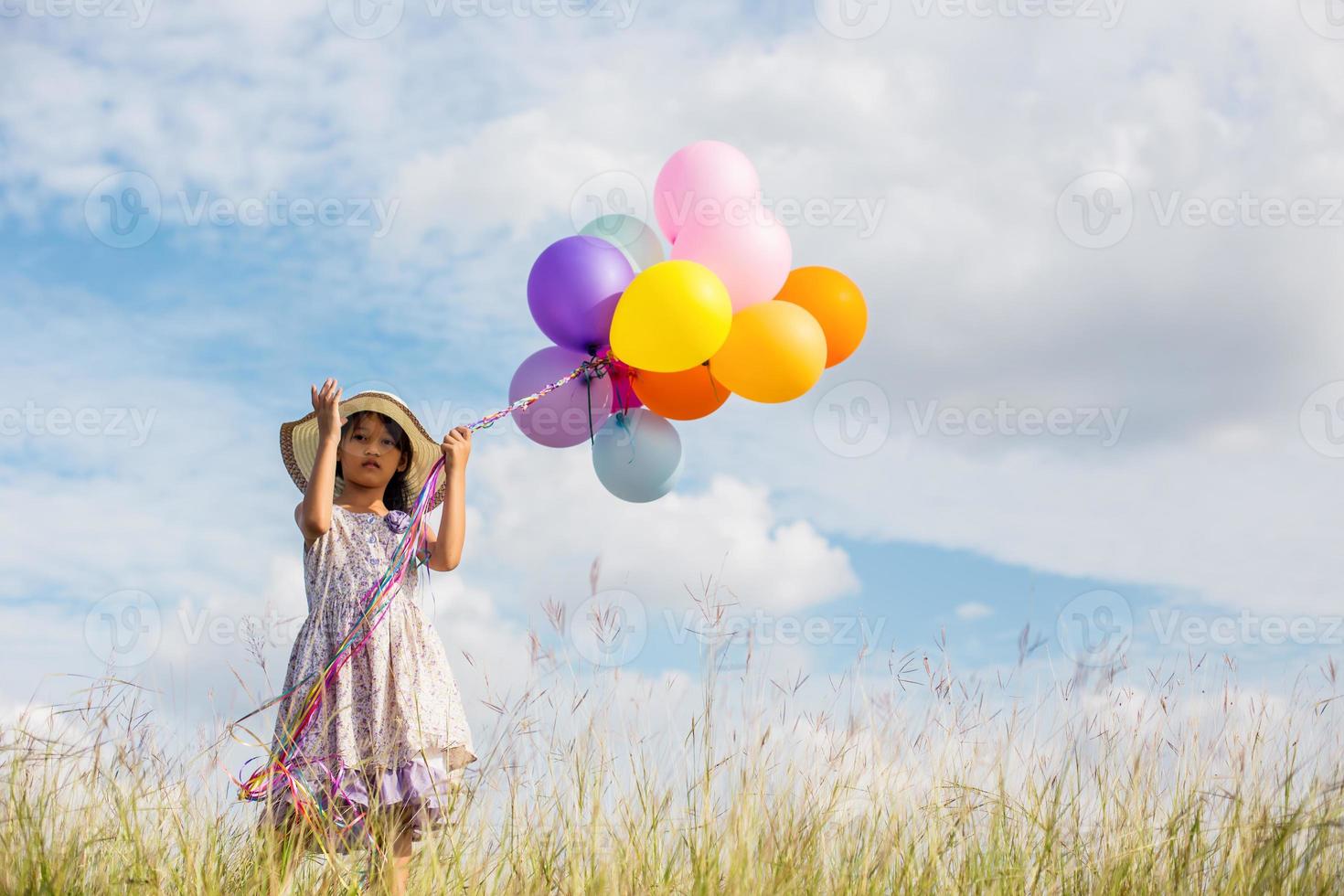 süßes kleines mädchen, das bunte luftballons auf der wiese gegen blauen himmel und wolken hält und die hände ausbreitet. foto