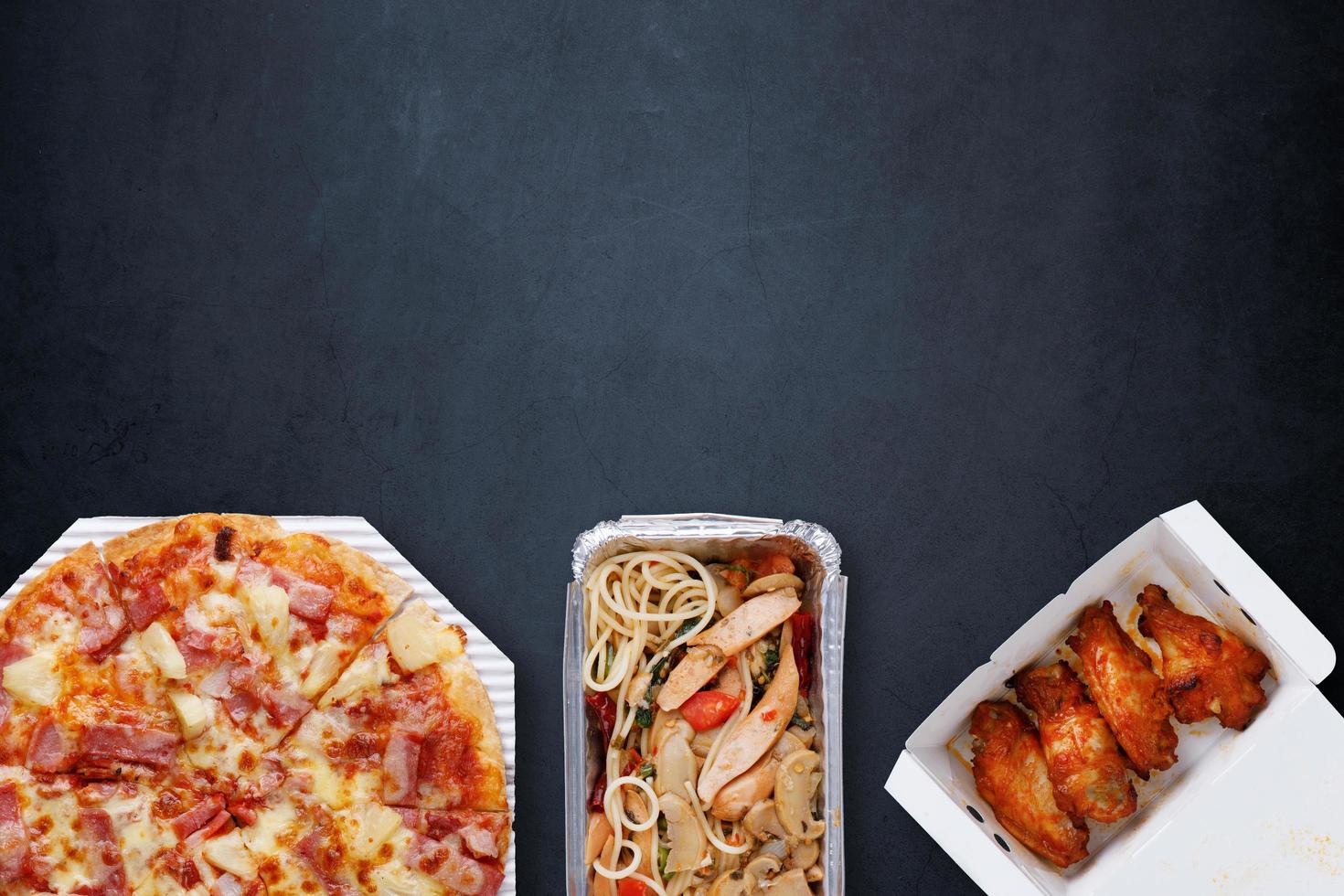 Lebensmittel-Lieferbox-Set. hawaiianische pizza mit dünner kruste auf einem papiertablett, gegrilltes hähnchen bbq in einer papierschachtel und spaghetti in einer hitzebeständigen schachtel auf einem grau-schwarzen beton mit kopierraum. Ansicht von oben. foto