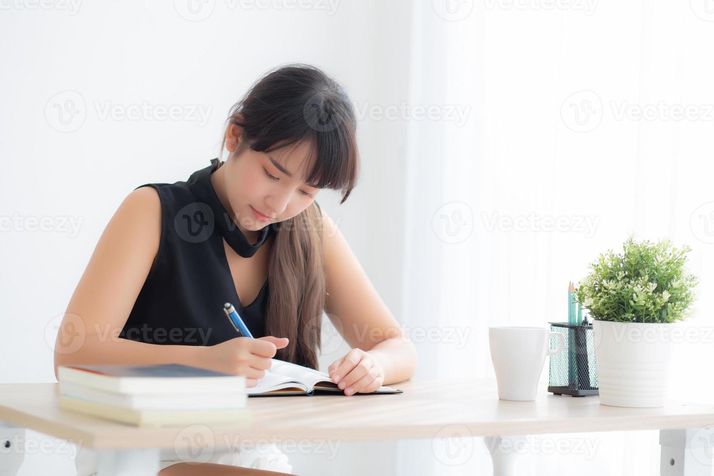 schöne junge asiatische frau, die lächelnd sitzend studiert und lernt, notizbuch und tagebuch im wohnzimmer zu hause zu schreiben, mädchenhausaufgaben, geschäftsfrau, die am tisch arbeitet, bildungskonzept. foto