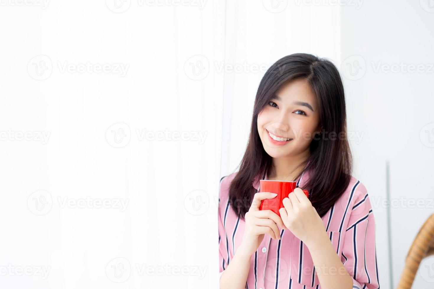 schöne junge asiatische frau des porträts mit trinken einer tasse kaffee stehendem vorhangfensterhintergrund im schlafzimmer, mädchen entspannen morgens zu hause, lebensstilkonzept. foto