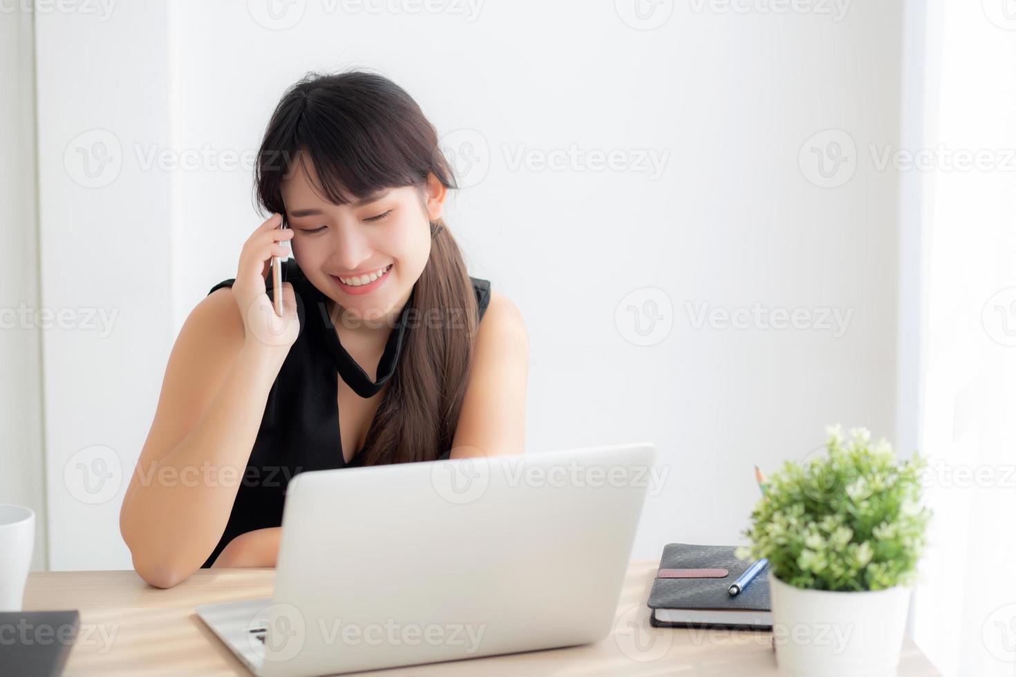 schöne junge freiberufliche asiatische frau, die lächelnd an einem laptop arbeitet und mit einem mobiltelefon im schreibtischbüro mit einem professionellen, mädchen, das ein notebook verwendet, einem business- und lifestyle-konzept arbeitet. foto