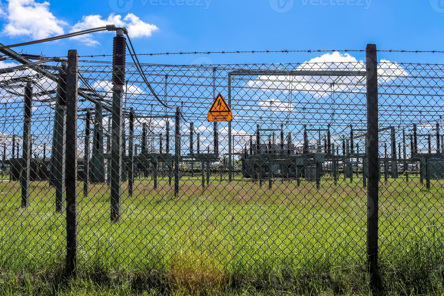 Schild mit den Worten Hochspannung in deutscher Sprache am Zaun eines großen Umspannwerks, das an einem sonnigen Tag elektrische Energie mit vielen Stromleitungen verteilt foto