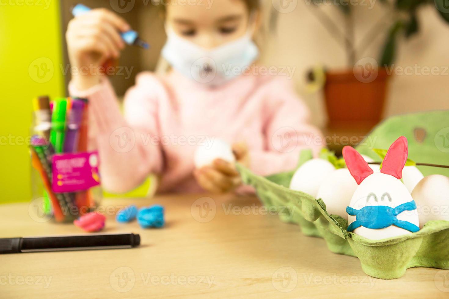 Ein Mädchen in den Ohren eines Hasen macht aus einem Ei und Plastilin einen Osterhasen in einer medizinischen Maske. diy sitzt zu hause, bereitet sich auf einen religiösen feiertag vor, inneneinrichtung. foto