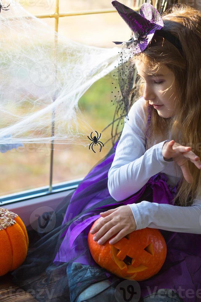 ein mädchen in einem hexenkostüm auf der fensterbank am fenster spielt mit einem spinnennetz und einer spinne und schmückt das haus für den halloween-urlaub. schreckliche Landschaften, Angst und Schrecken, Kürbisse foto