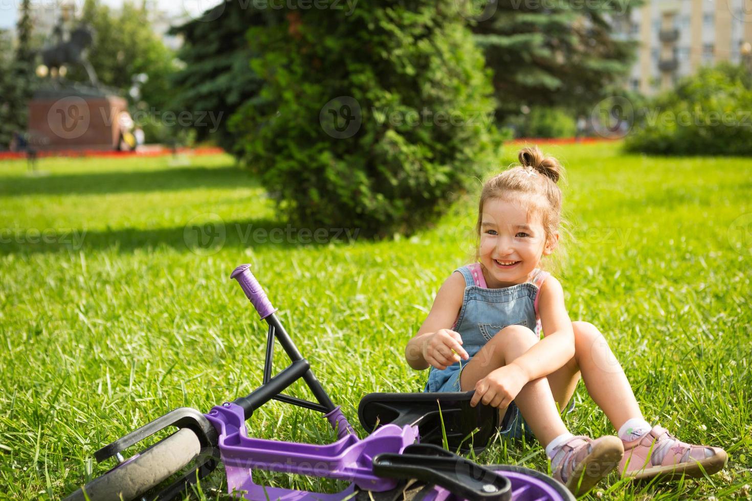 Fröhliches Mädchen, das im Sommer auf dem Gras in einem Park in der Nähe des lila Laufrads sitzt. aktive Unterhaltung für Kinder, Roller für Kleinkinder, glückliches Kind. Platz kopieren. sommer hintergrund foto