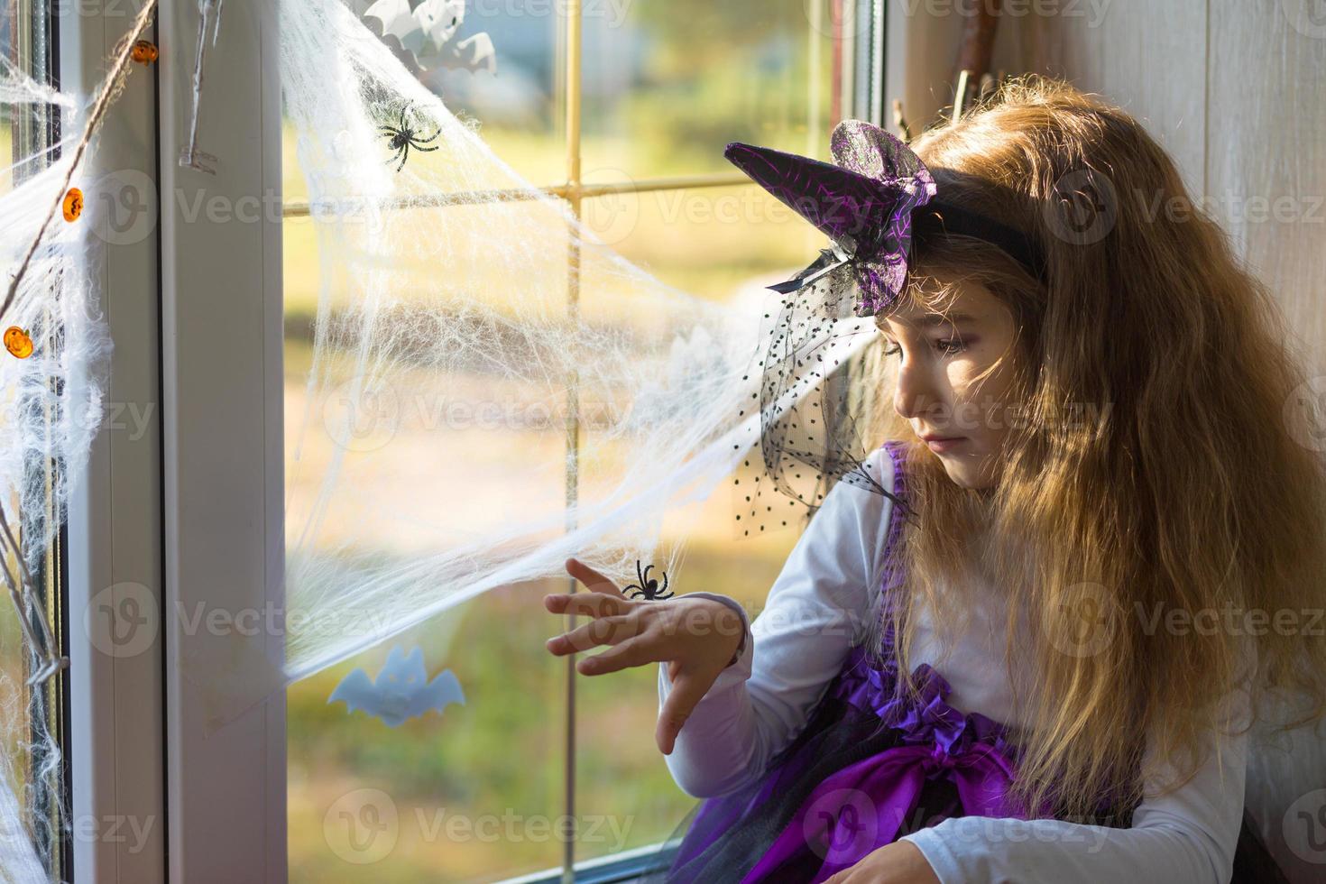 ein mädchen in einem hexenkostüm auf der fensterbank am fenster spielt mit einem spinnennetz und einer spinne und schmückt das haus für den halloween-urlaub. schreckliche Landschaften, Angst und Schrecken, Kürbisse foto