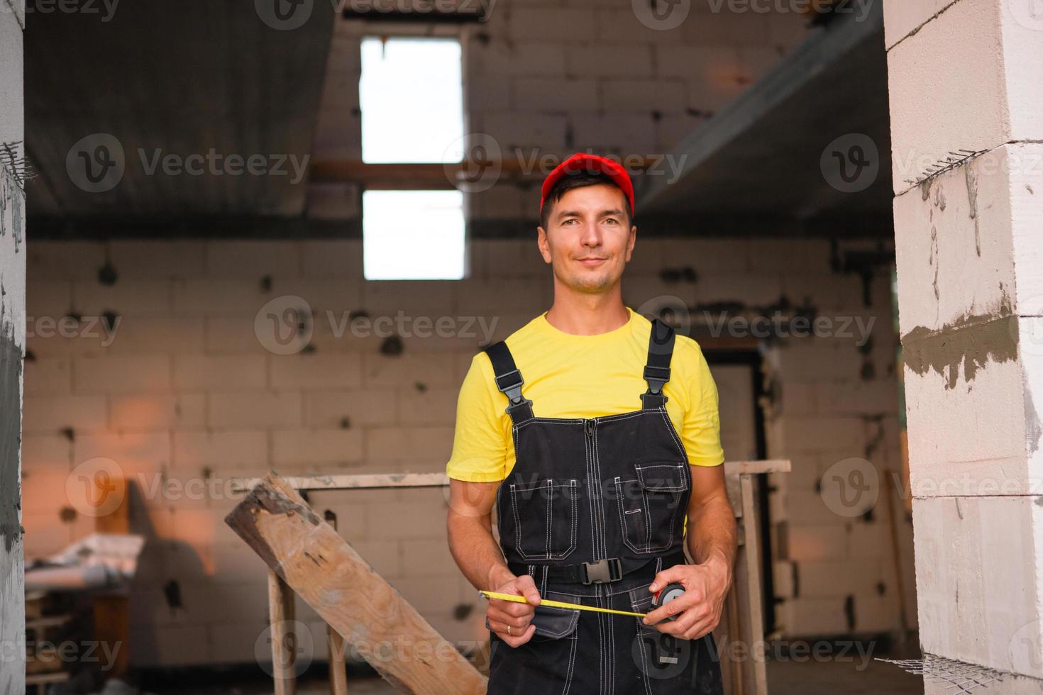 bauarbeiteringenieur auf der baustelle misst die länge der fensteröffnung und der ziegelwand mit einem maßband. Mann in Arbeitskleidung - Overall und Mütze foto