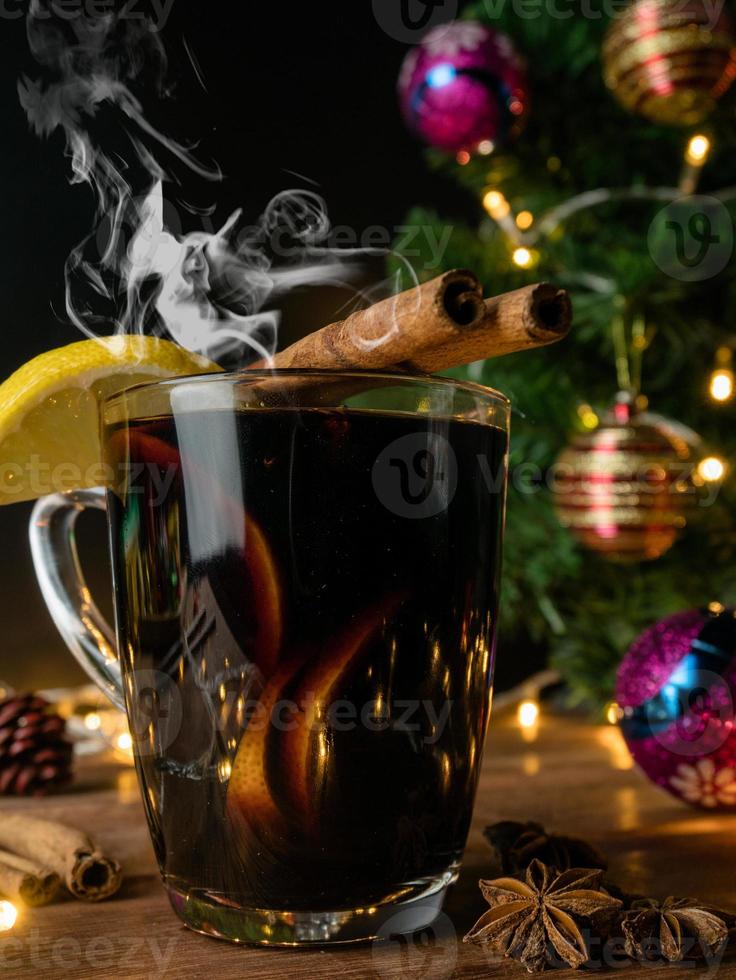 heißes getränk alkohol, rotwein in einer tasse glas gewürze zimtstange und zitrusfrüchte. dekorierte tanne festlich auf holztisch. feier weihnachten und frohes neues jahr konzept. foto