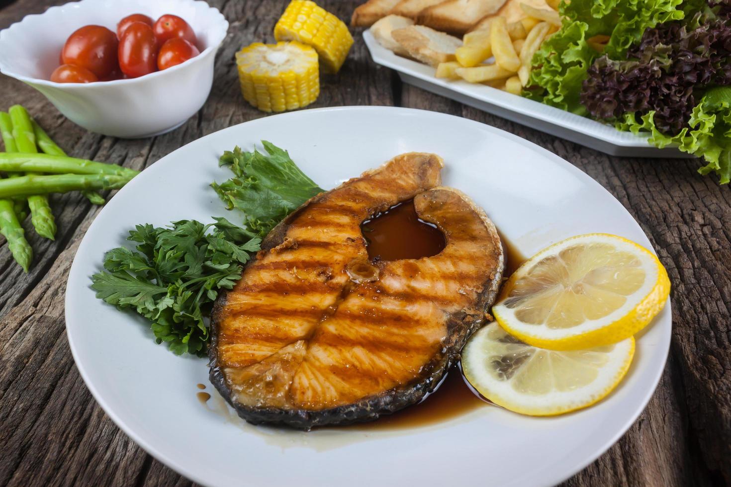 Lachssteak mit Soße auf einem weißen Teller mit Zitrone auf dem Teller und knusprigen Pommes Frites. Viele Gemüsesorten werden um die Schüssel herum auf dem Holzboden platziert. foto