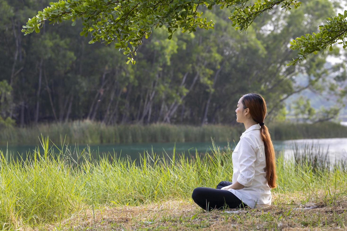 Frau, die entspannt im öffentlichen Park sitzt und Meditation praktiziert, um im Sommer Glück aus innerer Friedensweisheit unter dem Baum zu erlangen foto