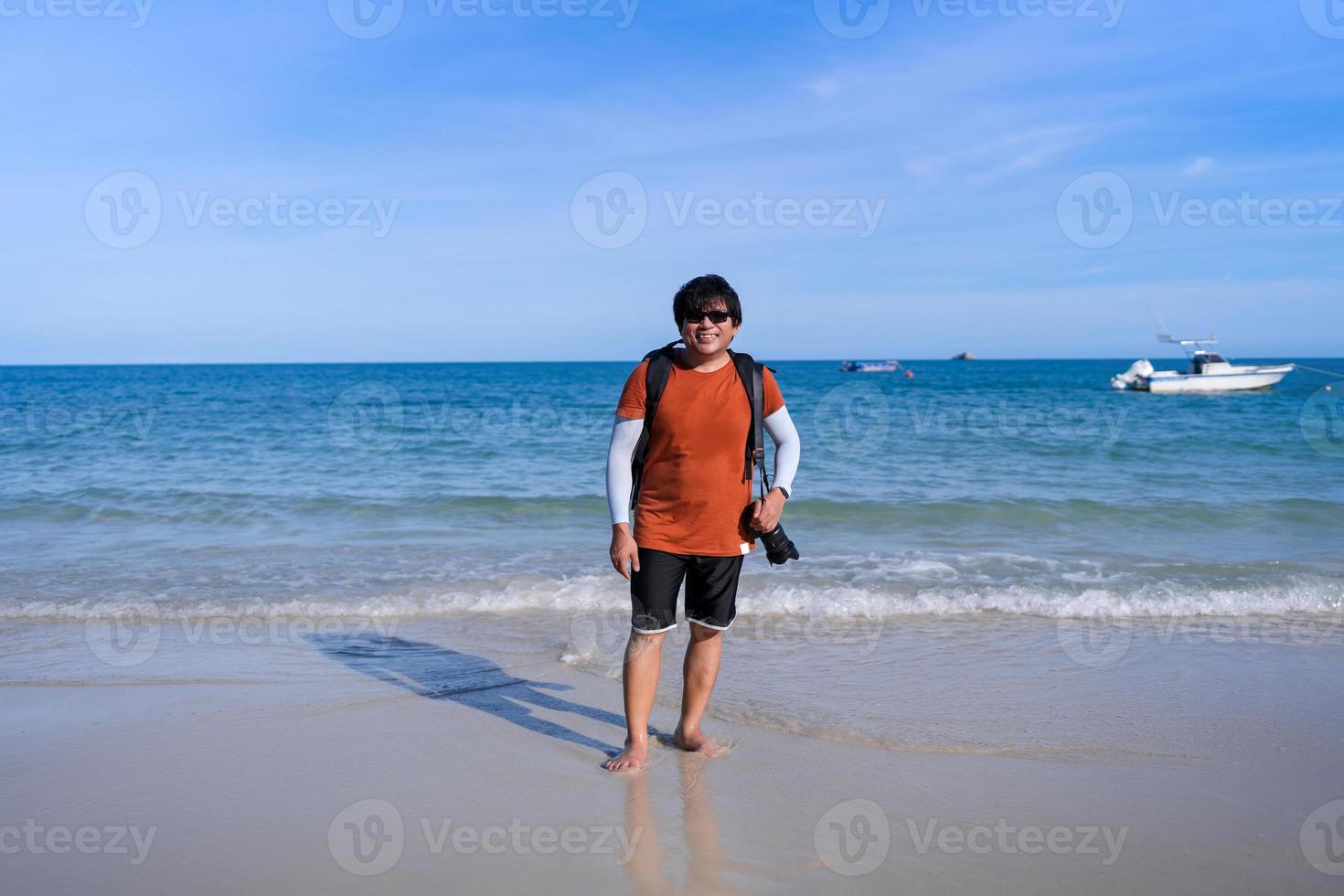 glücklicher asiatischer rucksackfotograf touristischer mann mittleren alters, der am strand steht und lächelt foto