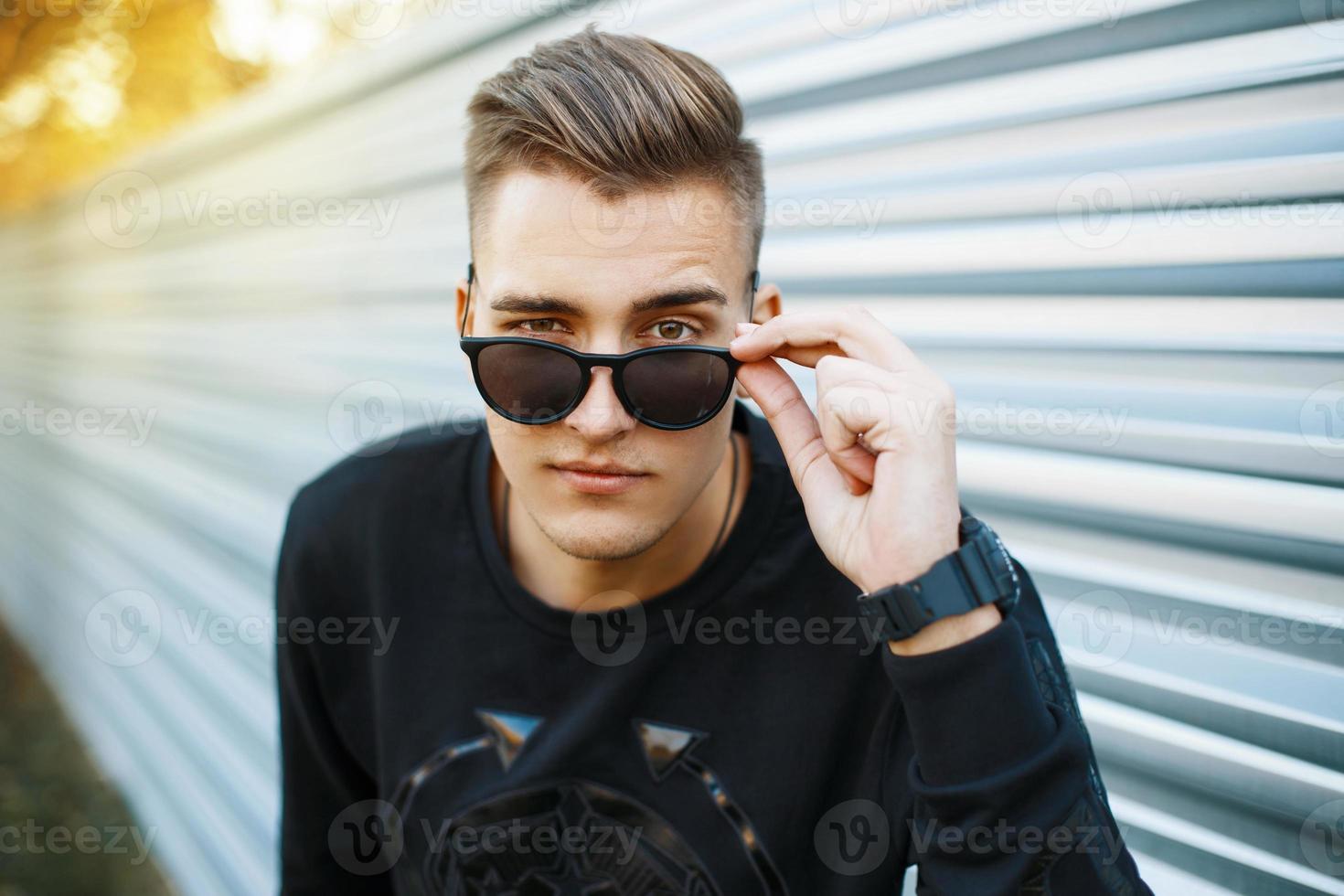 Junger gutaussehender Mann in stylischer schwarzer Kleidung, der durch eine Sonnenbrille schaut. foto