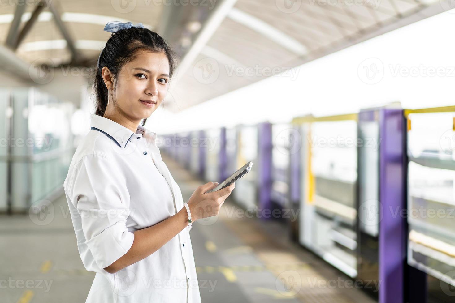 asiatische Passagierin mit lässigem Anzug, die das Smartphone benutzt, während sie am Bahnhof auf den Skytrain oder die U-Bahn wartet, um in der Großstadt, im Lifestyle- und Transportkonzept zu reisen foto