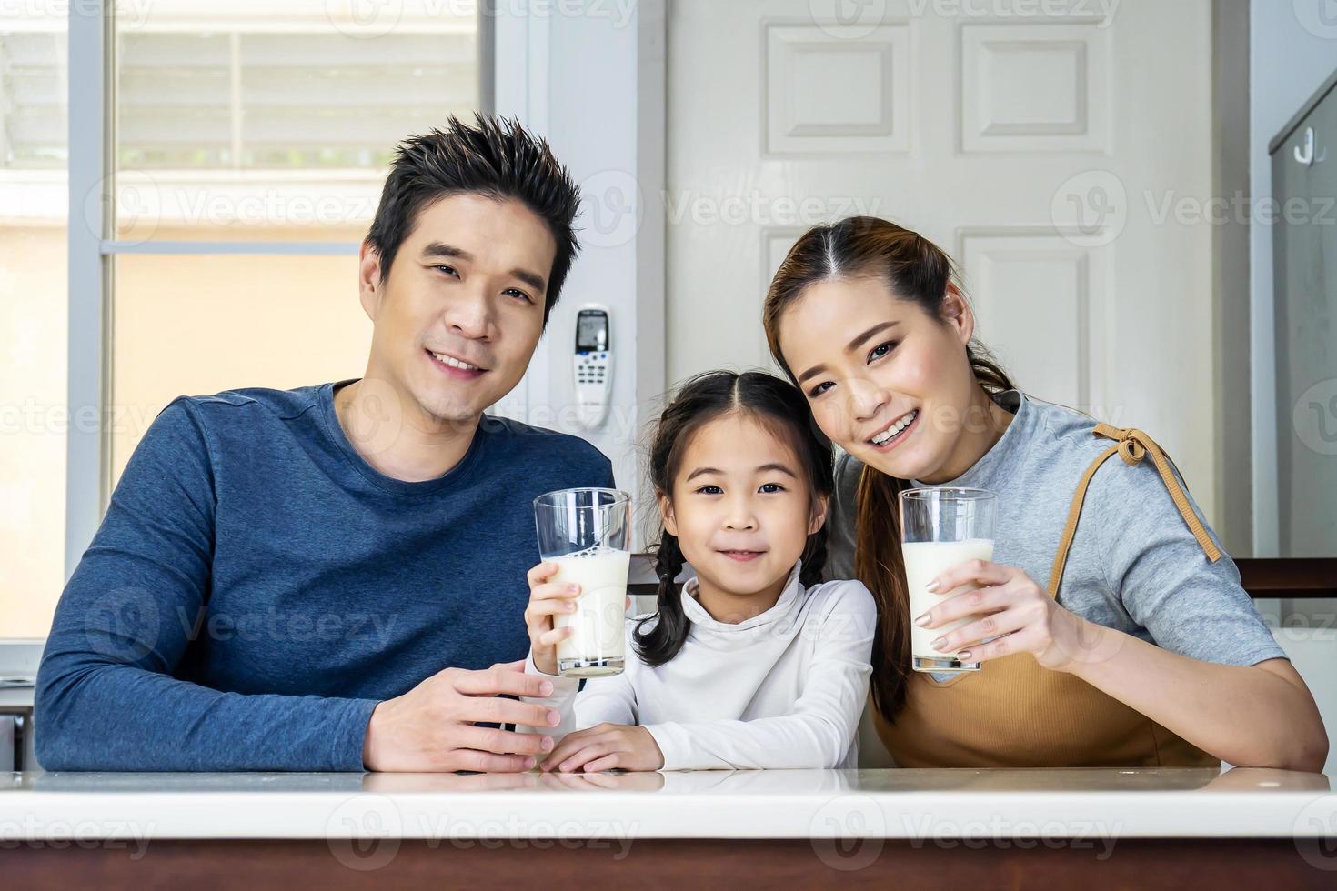 glückliche familie, die spaß in der küche hat. asiatischer vater, mutter und kleine tochter verbringen zeit zusammen und frühstücken, trinken und halten gläser milch am tisch foto