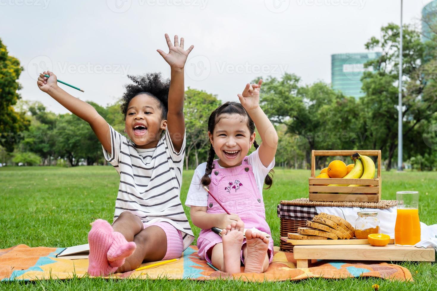 glückliche kindheit, zwei afroamerikanische kleine kinder, die mit bleistift auf notizbuch zeichnen, während sie auf der matte im freien liegen, zwei kleine mädchen, die spaß haben, hoben glücklich die hände, nachdem sie die hausaufgaben beendet hatten foto