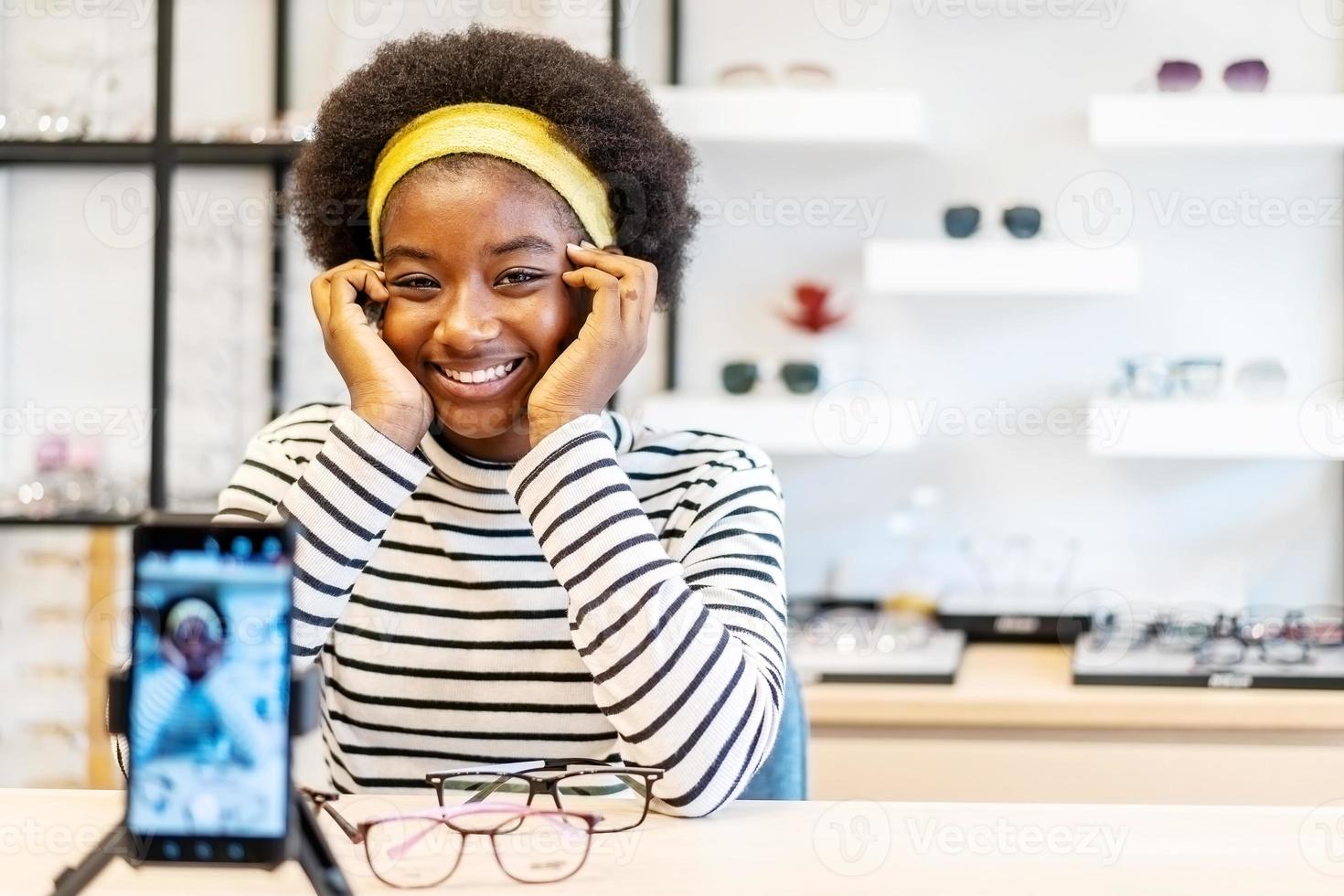 junge afroamerikanische unternehmerin ist mode-vloggerin, die brille auf handy zeigt und lächelt, sie online live-streaming im brillengeschäft zu verkaufen foto
