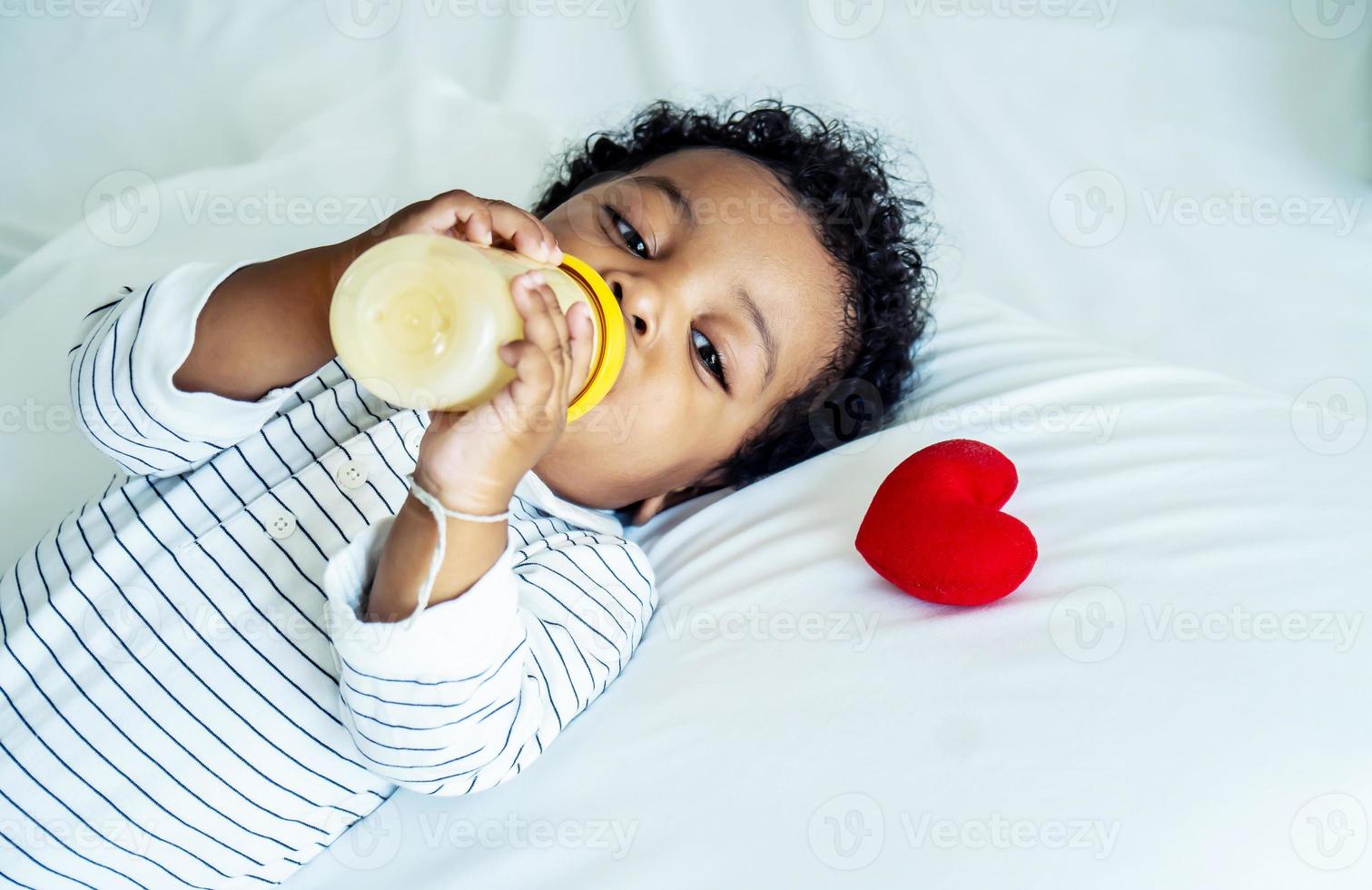 afrikanisch-amerikanischer süßer Junge, der Milch aus der Flasche trinkt und zu Hause mit einem roten Herzen auf dem Bett schläft, Kinder brauchen Liebe und Fürsorge foto