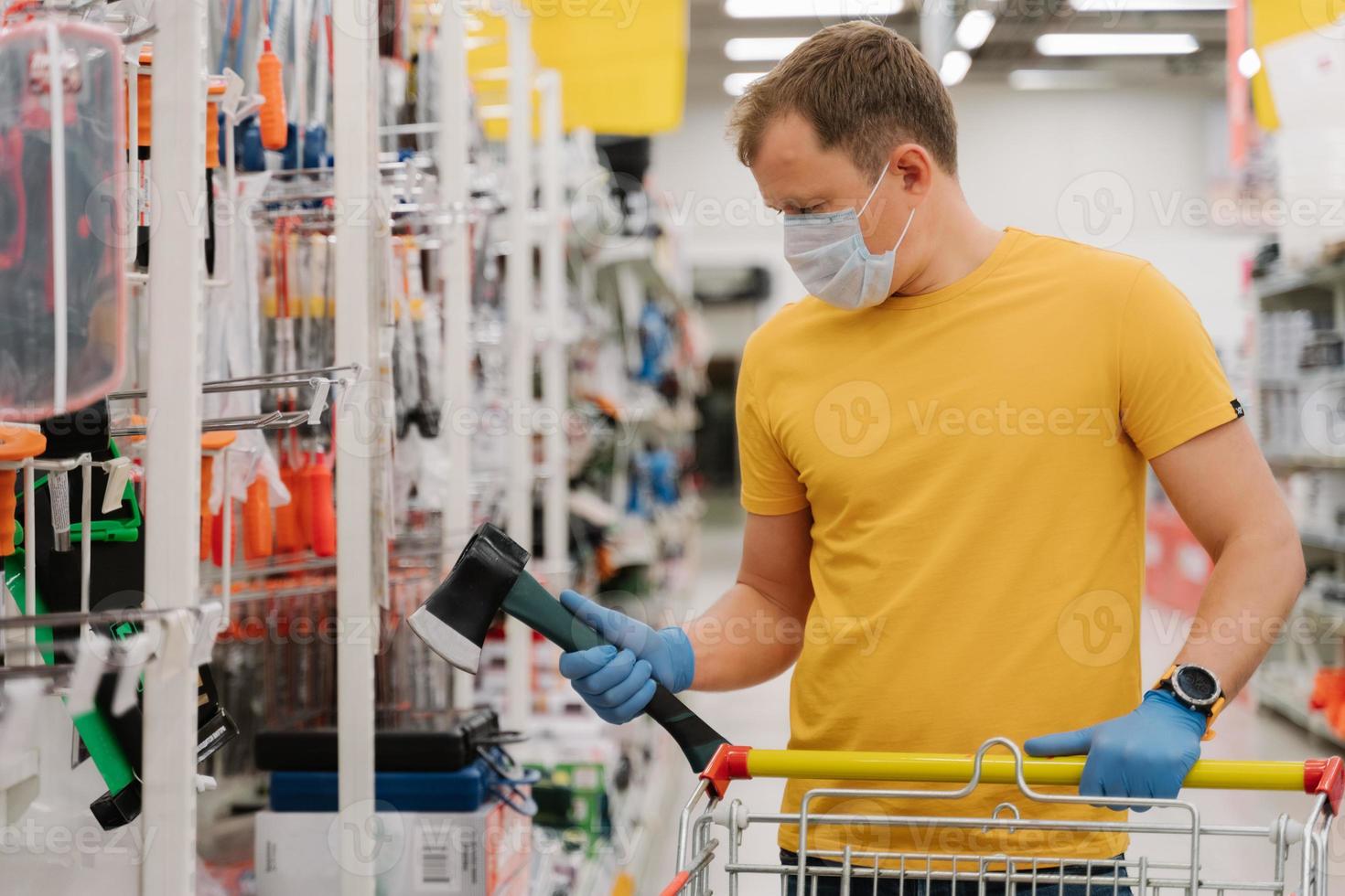 Schutz vor dem Coronavirus. mann wählt axt in der bauabteilung des einkaufszentrums, posiert mit einkaufswagen, trägt einweggummihandschuhe und maske, verhindert die ausbreitung von covid-19. Pandemie Ausbruch foto