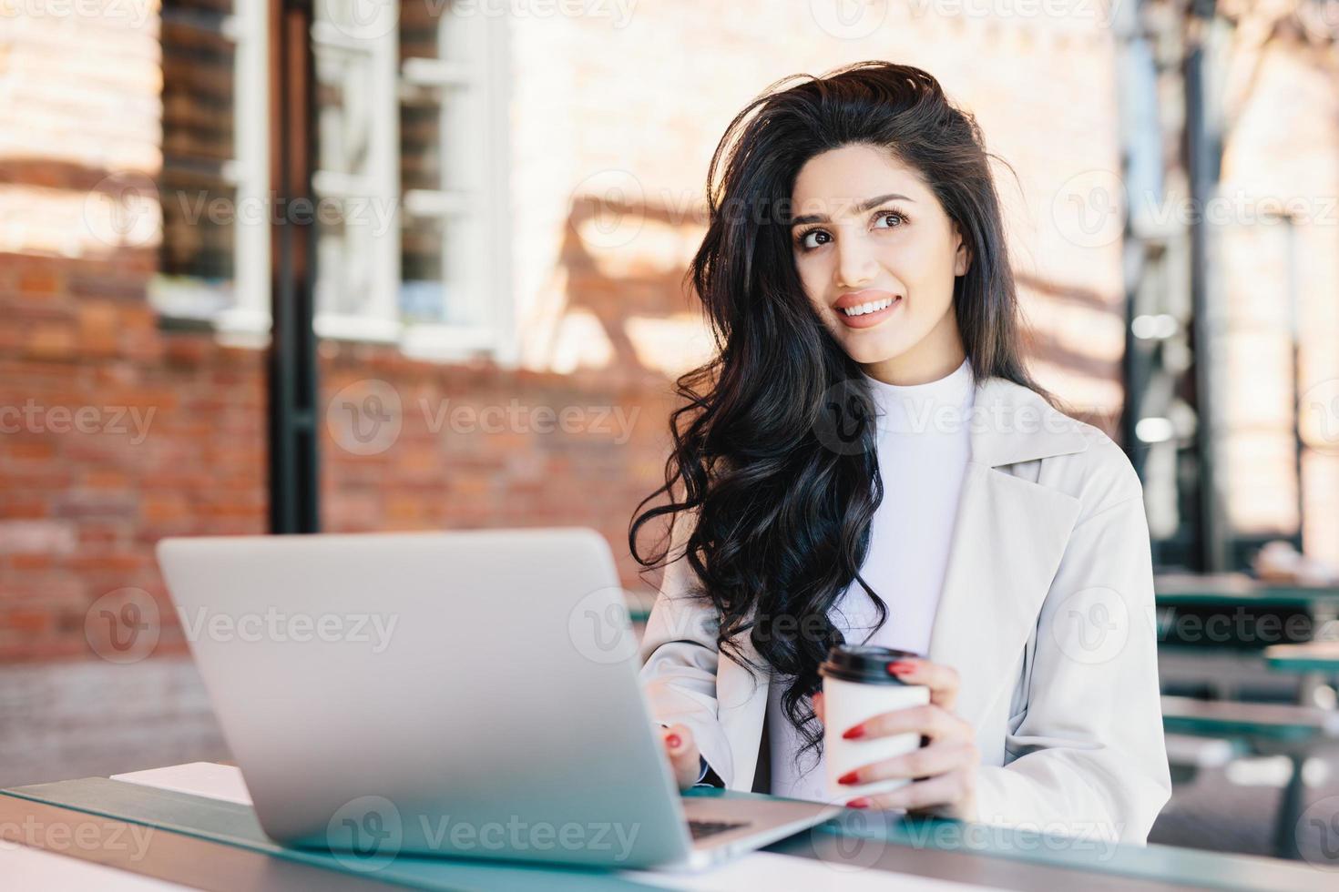 Modische brünette Frau mit langen, luxuriösen Haaren, elegant gekleidet und mit nachdenklichem Ausdruck in die Ferne blickend, die von Urlaub träumt, während sie am Laptop im Café im Freien arbeitet. Technologiekonzept foto