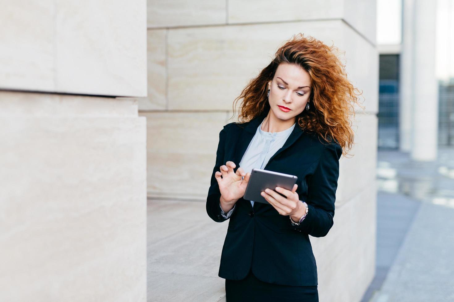elegante Dame mit lockigem Haar, schwarzem Anzug, Nachrichten eingeben oder Geschäftsbericht erstellen, während sie einen Tablet-Computer verwendet. Unternehmerin, die an neuen Geschäftsprojekten arbeitet. Geschäft und Karriere foto