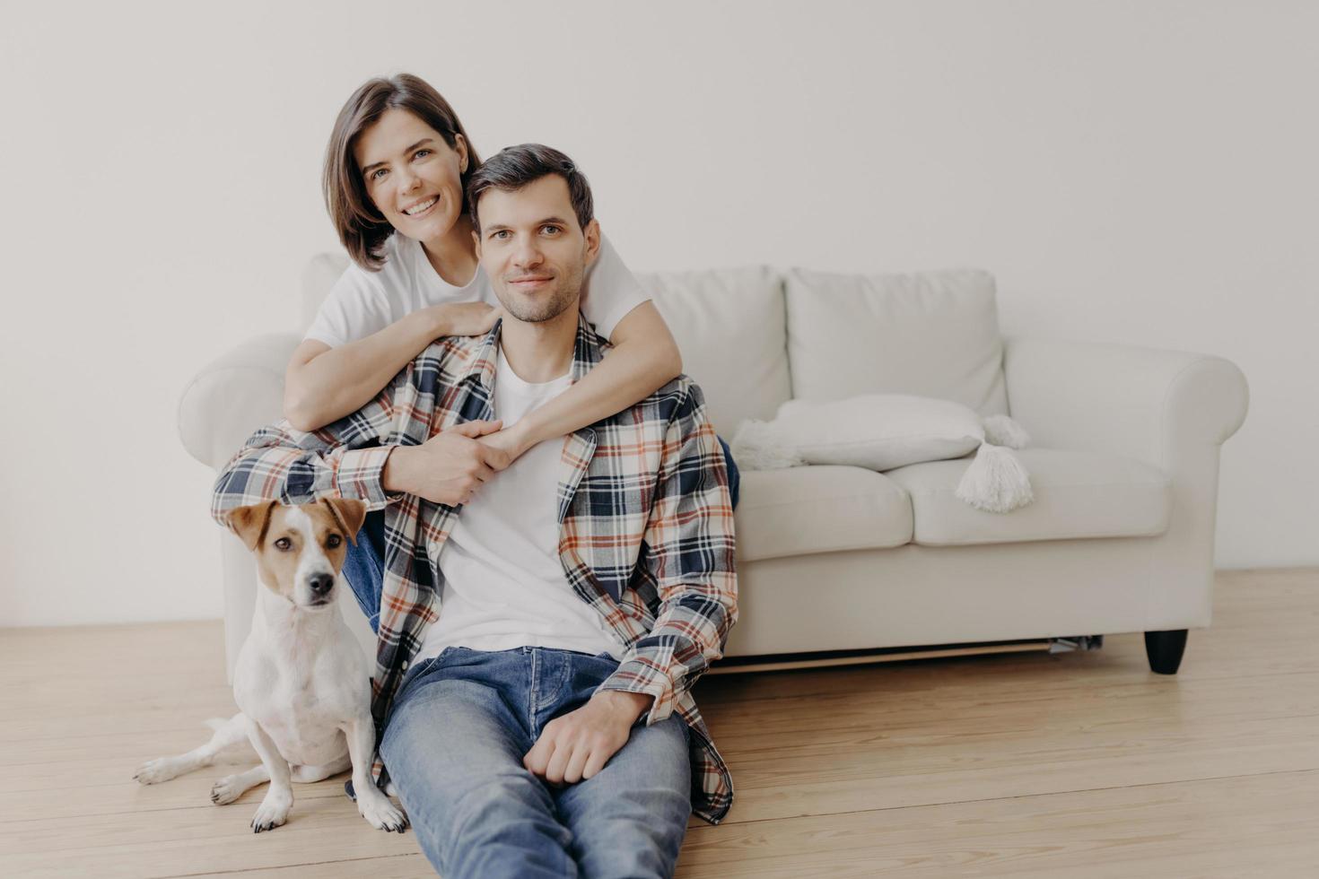 Foto einer glücklichen Frau umarmt ihren Mann, posiert auf dem Sofa, kleiner Hund sitzt auf dem Boden neben dem Besitzer, schaut alle mit glücklichen Gesichtsausdrücken in die Kamera, hat eine romantische Beziehung. Familienporträt. Menschen und Heimat