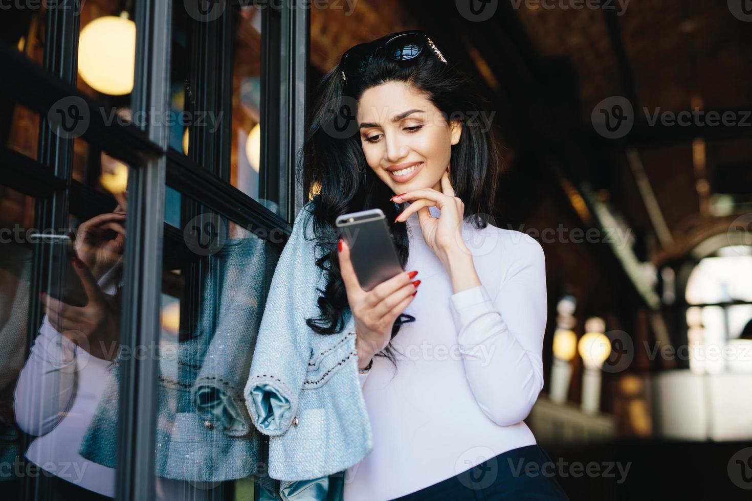 wunderschöne brünette frau mit sonnenbrille, die formell mit dem handy gekleidet ist, flirtet mit einem gutaussehenden mann im internet, wartet auf seine antwort, schaut mit vergnügen und großer freude auf den bildschirm im café foto