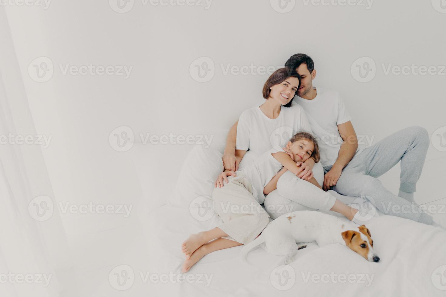 Zufriedener Ehemann, Ehefrau und ihre kleine Tochter posieren in einem geräumigen Schlafzimmer mit Lieblingshaustier, haben Spaß, umarmen und drücken Liebe zueinander aus, Unterstützer in schwierigen Situationen. Familienzeitkonzept foto