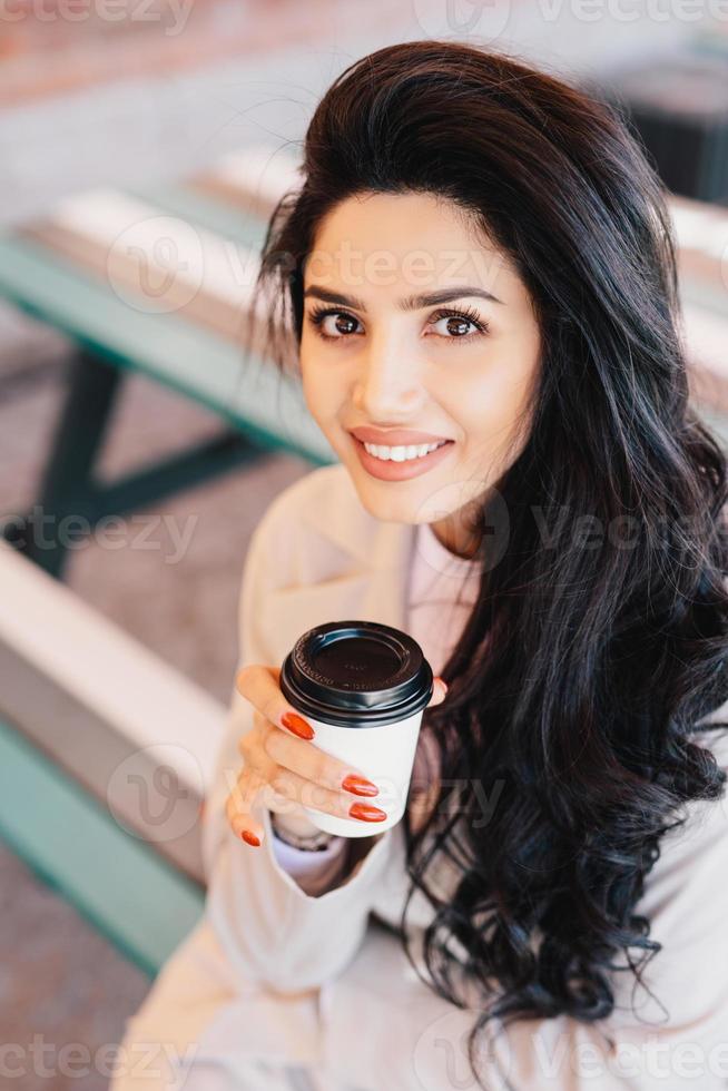 Außenaufnahme einer schönen brünetten Frau mit wunderbarem Aussehen, die weiße Kleidung trägt und eine Papptasse mit köstlichem Kaffee hält. Kaukasische Frau mit angenehmem Lächeln, die ihre Freizeit im Freien verbringt foto