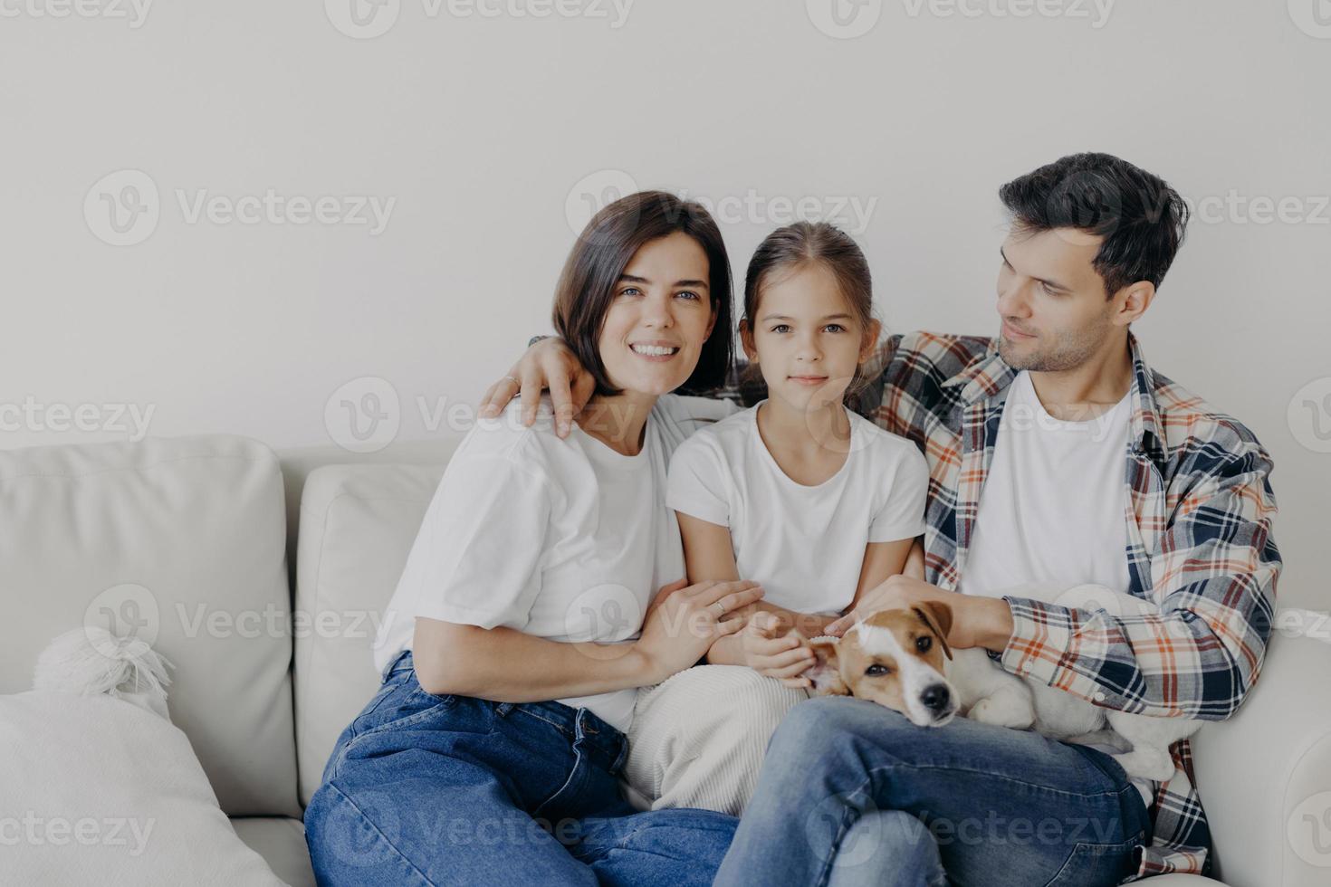 Familienzeitkonzept. Fröhliche Mutter, Vater und kleines Mädchen umarmen sich alle zusammen, verbringen Freizeit im Wohnzimmer ihres neuen Zuhauses, sitzen auf dem Sofa, spielen mit dem Hund, haben perfekte Beziehungen, genießen Komfort foto