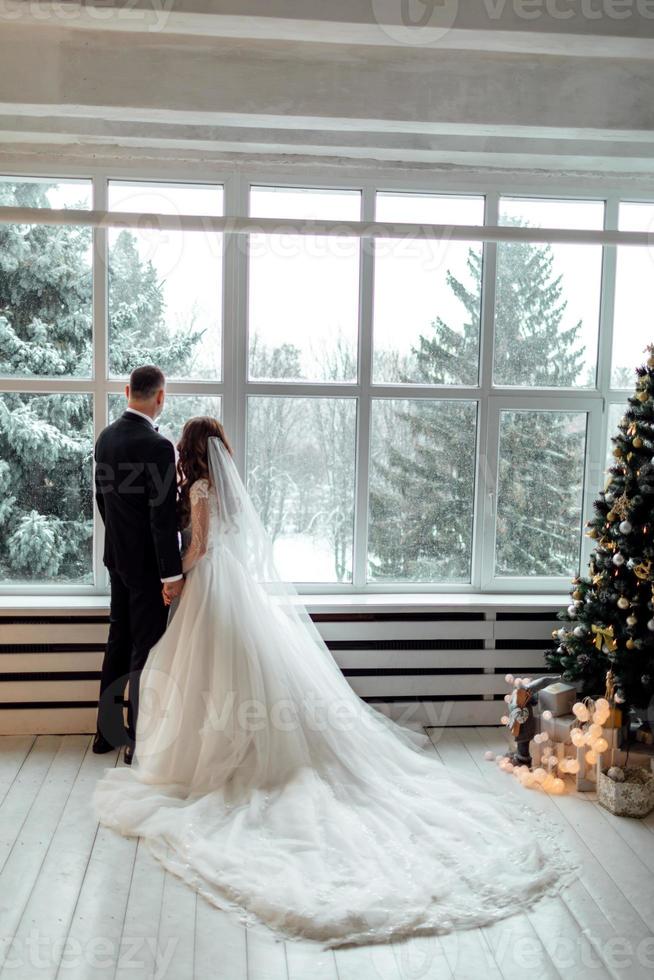 junges verliebtes paar braut und bräutigam posieren im studio auf hintergrund, der an ihrem hochzeitstag zu weihnachten in der nähe des großen panoramafensters mit weihnachtsbaum geschmückt ist. foto