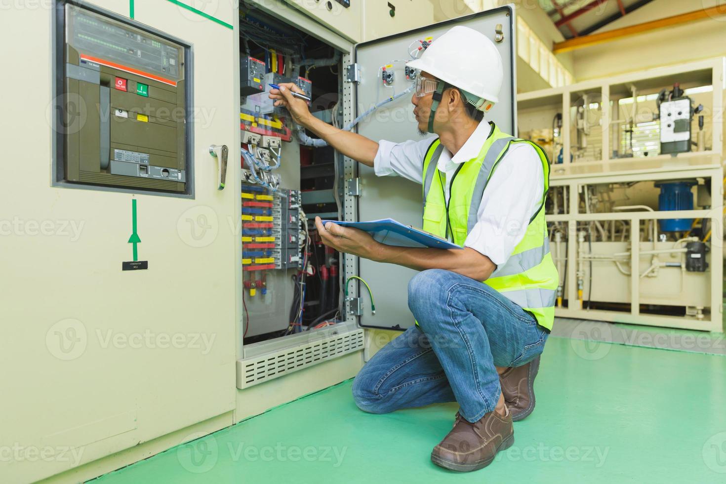 Ein behelmter Techniker hält ein Klemmbrett, während er den Elektroschaltschrank überprüft und Notizen macht. foto