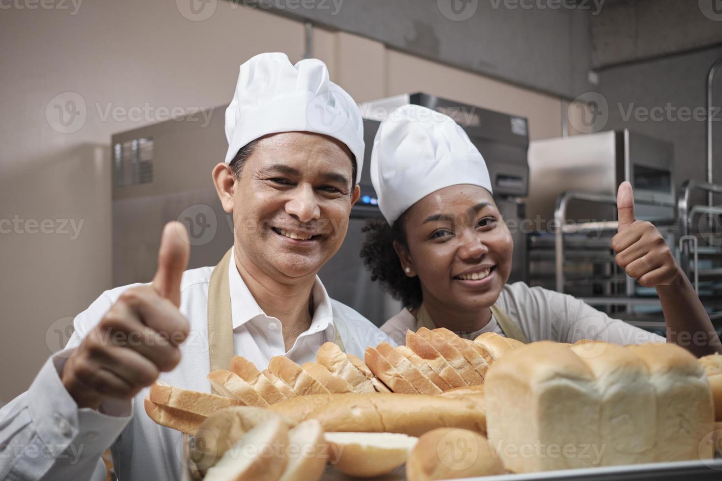 Porträt professioneller Köche in weißer Uniform, die mit fröhlichem Lächeln und Daumen nach oben mit einem Tablett Brot in der Küche in die Kamera blicken. Freund und Partner von Backwaren und täglich frischer Bäckertätigkeit. foto