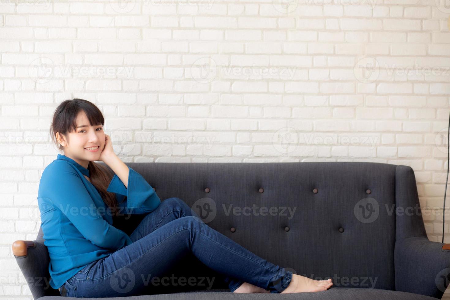 schöne junge asiatische frau des porträts, die glücklich sitzt und lächelt und kamera auf dem sofa mit lässigem wohnzimmer betrachtet, mädchen fröhlich und entspannt auf der couch zu hause, lebensstilkonzept. foto