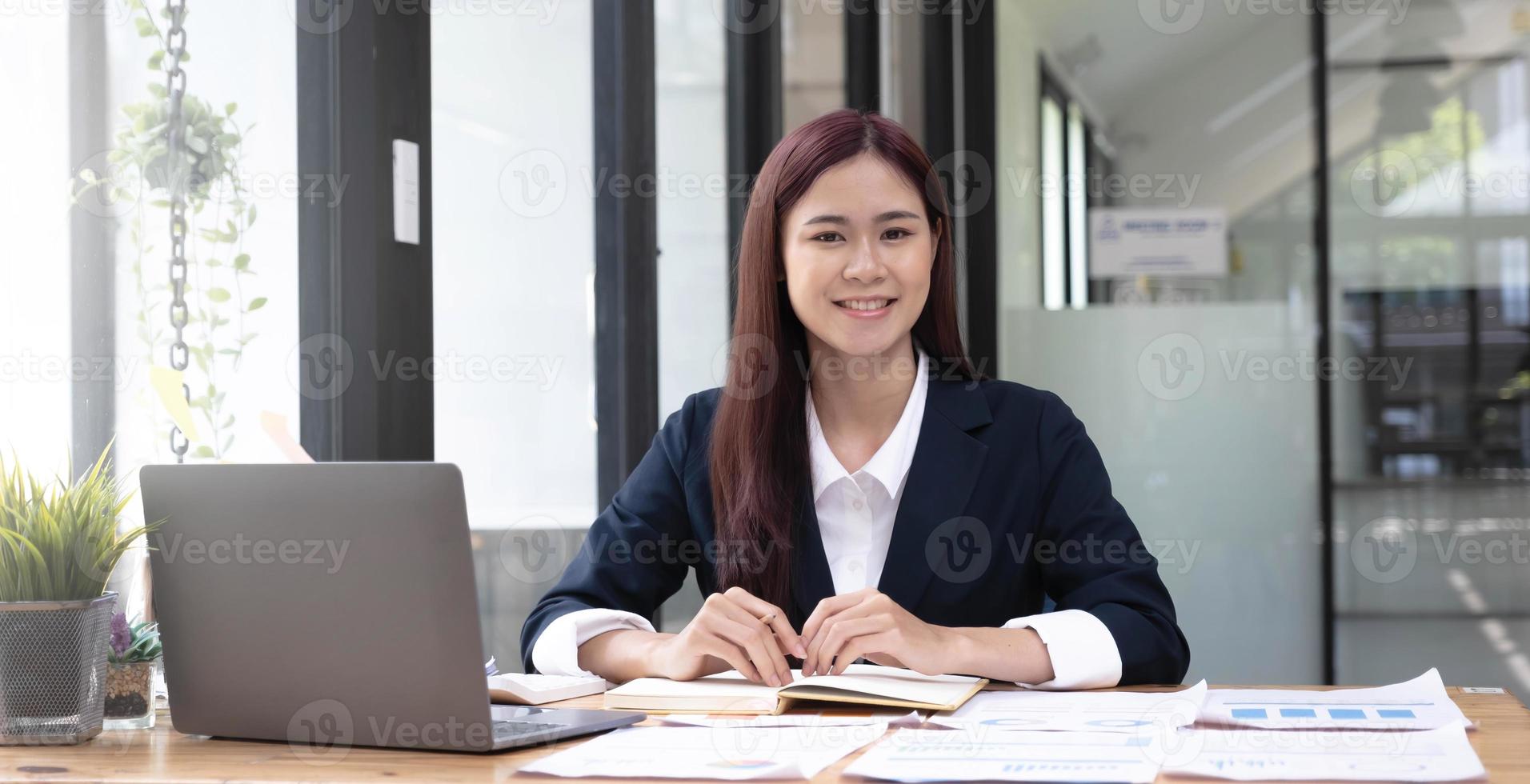 charmante asiatische frau, die im büro arbeitet und einen laptop verwendet, der in die kamera schaut. foto