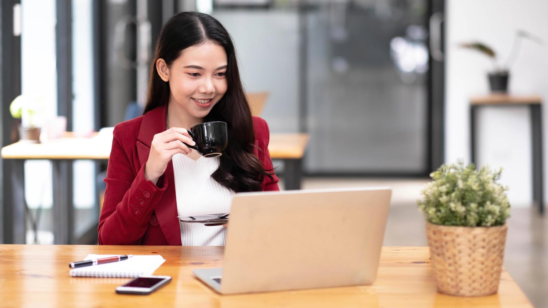 junge asiatische geschäftsfrau hat das vergnügen, die arbeit auf ihrem tablet und ihrem lieblingskaffee im büro zu beobachten. foto