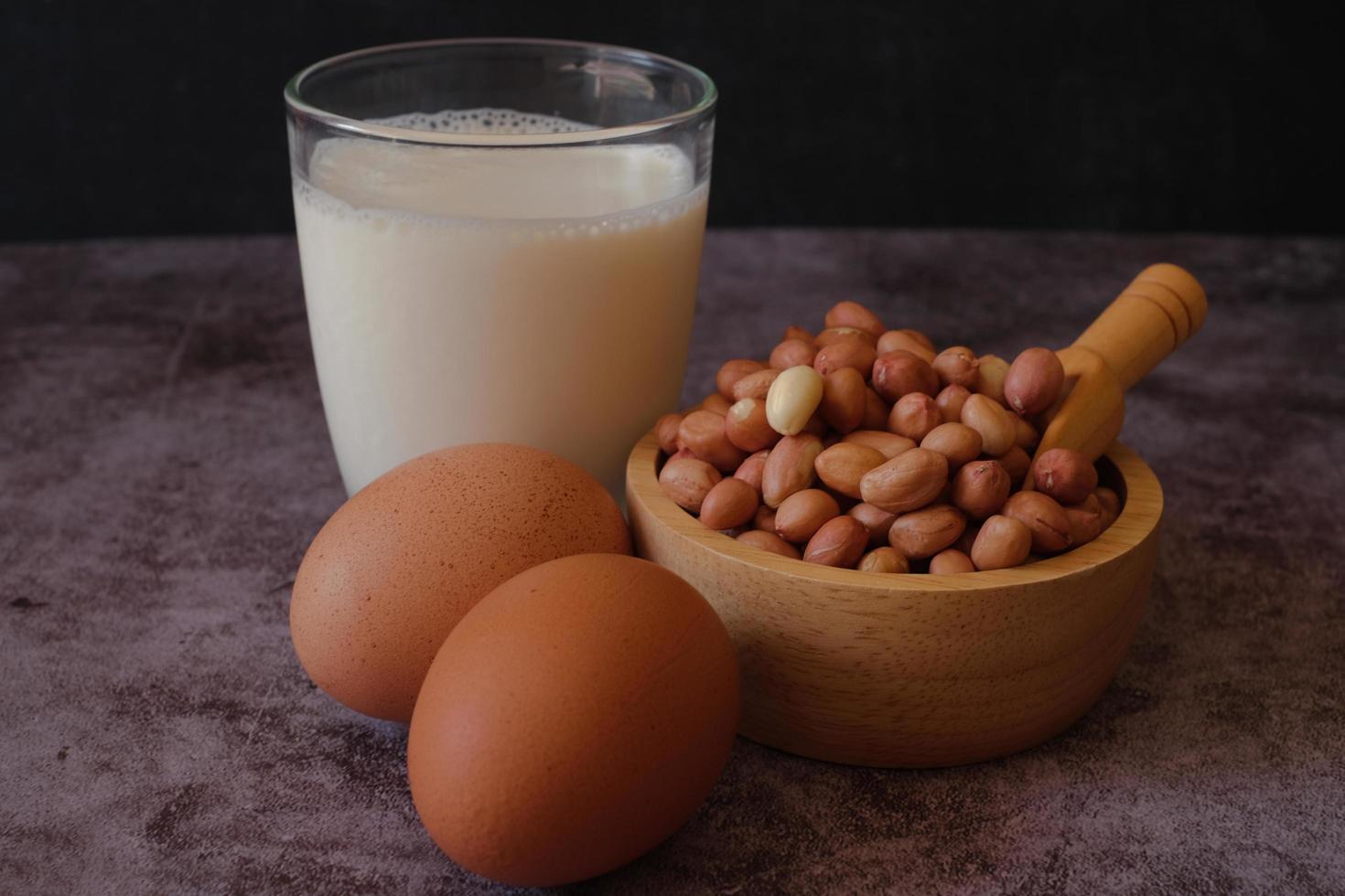 Protein-Lebensmittelgruppe. Rohe Erdnüsse in der Holzschale, Hühnerei und ein Glas Milch auf Zementhintergrund. gesundes Essen. foto