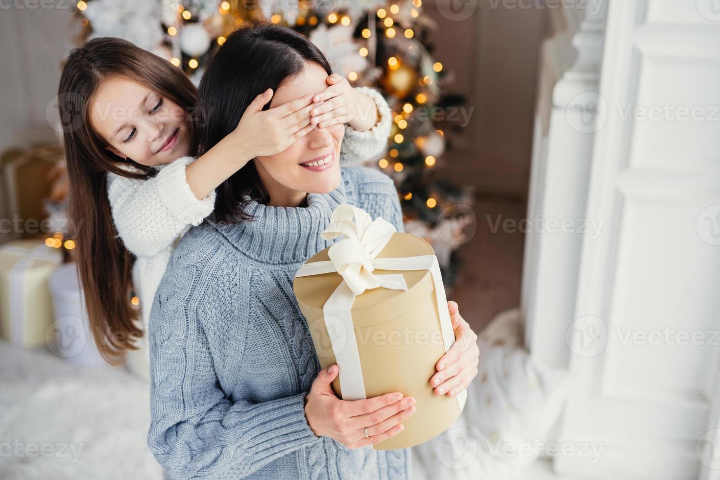 Kleines entzückendes weibliches Kind bereitet Überraschung für ihre Mutter vor, schließt Augen und gibt verpacktes Geschenk als Stand am Neujahrshintergrund. hübsche Frau erhält Geschenk von Tochter. Überraschungskonzept foto