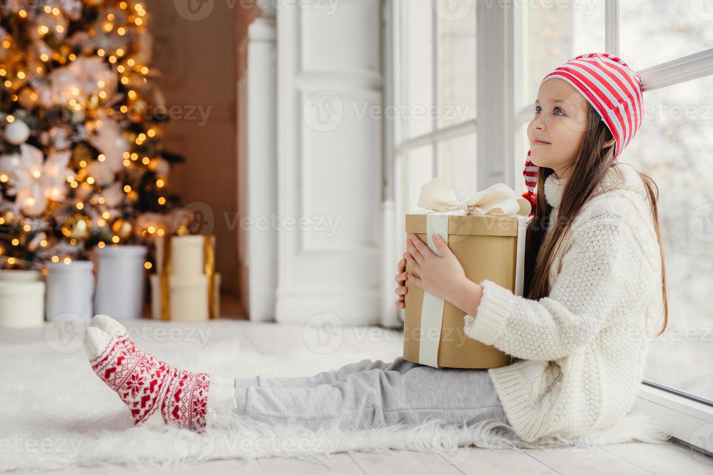 seitliches porträt eines ruhigen weiblichen kindes trägt einen weißen pullover, eine hose und warme socken, umarmt ein verpacktes geschenk, sitzt in einem gemütlichen zimmer auf dem boden und bewundert den neujahrsbaum. kinder- und ferienkonzept foto