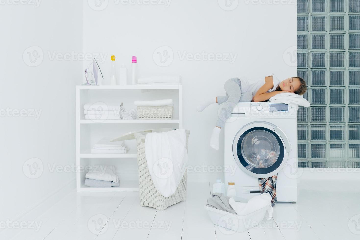 Vorschulkind schläft auf Waschmaschine, müde vom Waschen, posiert in weißer großer Waschküche mit Korb und Becken voller schmutziger Kleiderflaschen mit flüssigem Pulver. Kindheit, Hausarbeit foto