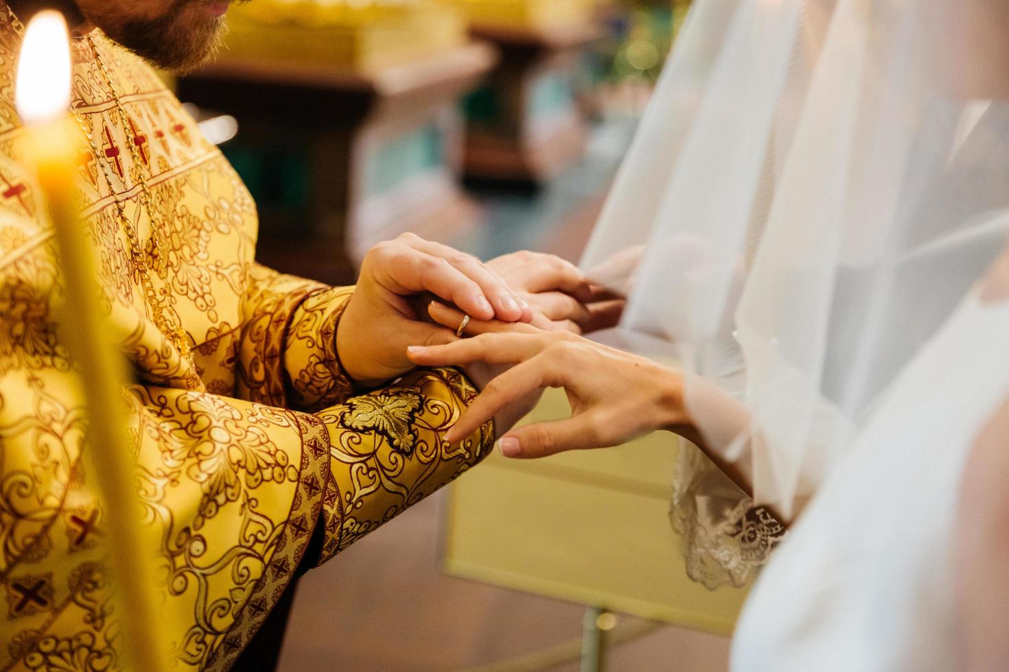 Ansicht des Priesters, der den Ring an der Hand der Braut anlegt, Pose in der christlichen Kirche während der Hochzeitszeremonie, brennende Kerze im Vordergrund foto