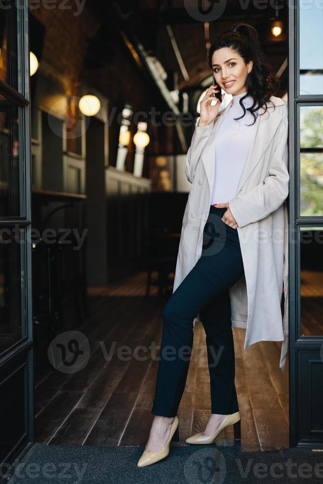 Stilvolle junge Frau mit dunklem Haar, weißem Mantel, schwarzer Hose und Schuhen mit Absätzen, die in die Kamera posieren, während sie vor Café-Hintergrund stehen und über das Telefon chatten und die Hand in der Tasche halten foto